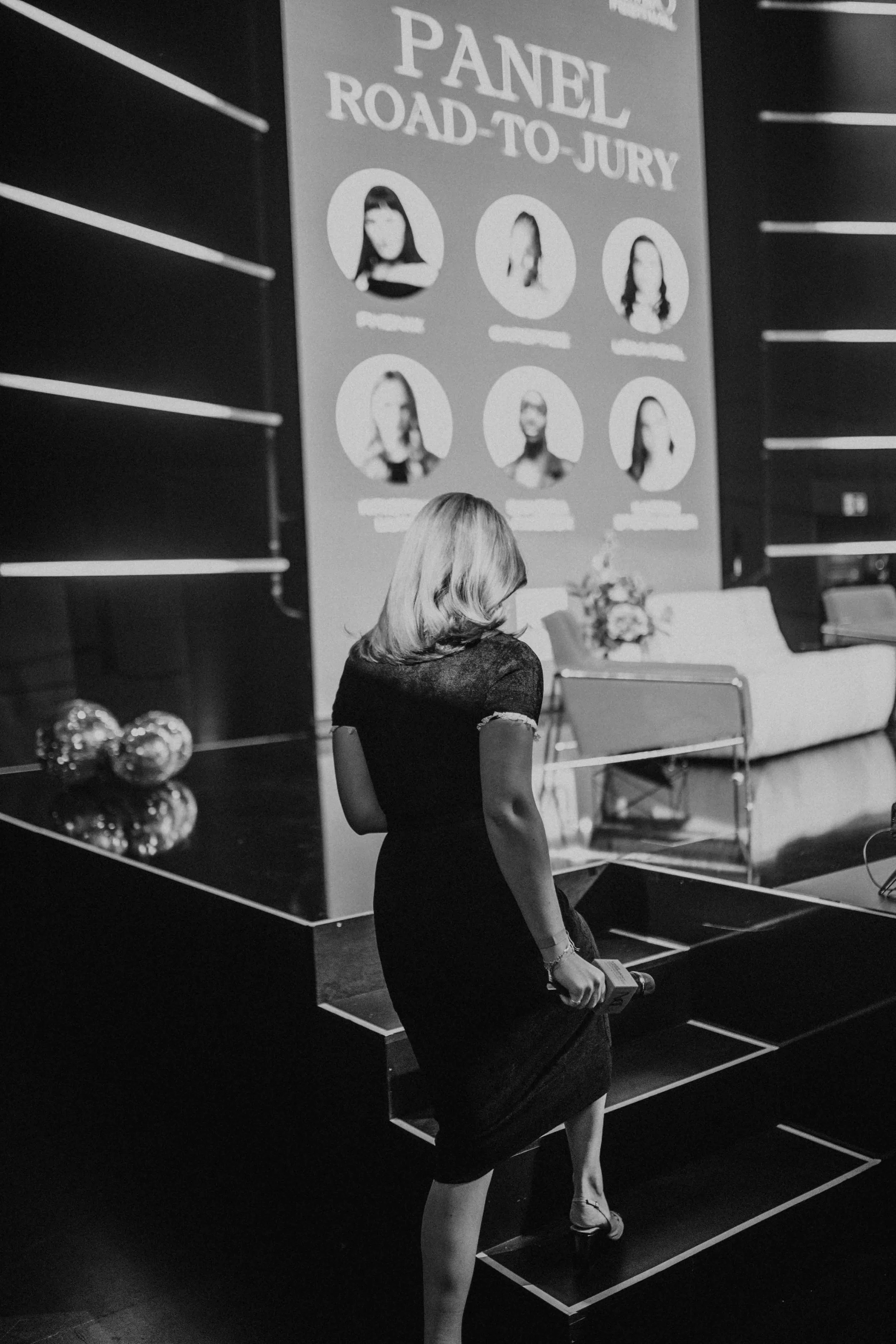 Frau im schwarzen Kleid vor einer Eventwand mit Porträtfotos