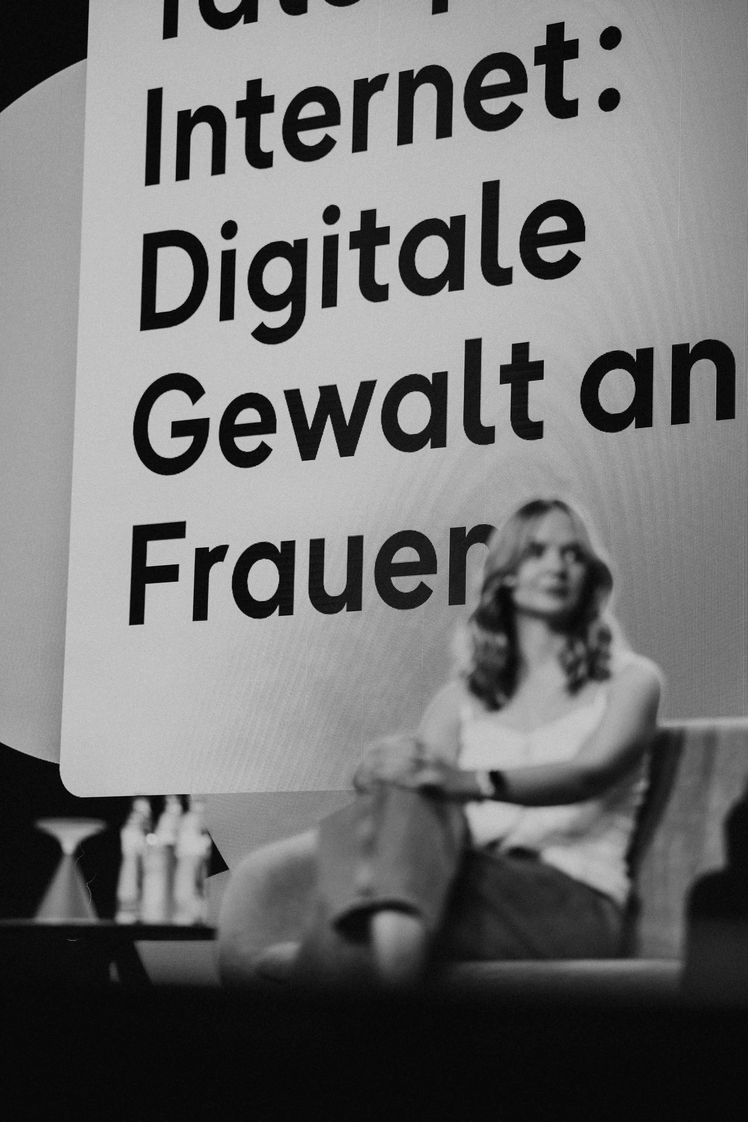 Schwarz-weißes Foto einer Frau auf einer Bühne vor der Aufschrift "Internet: Digitale Gewalt an Frauen"