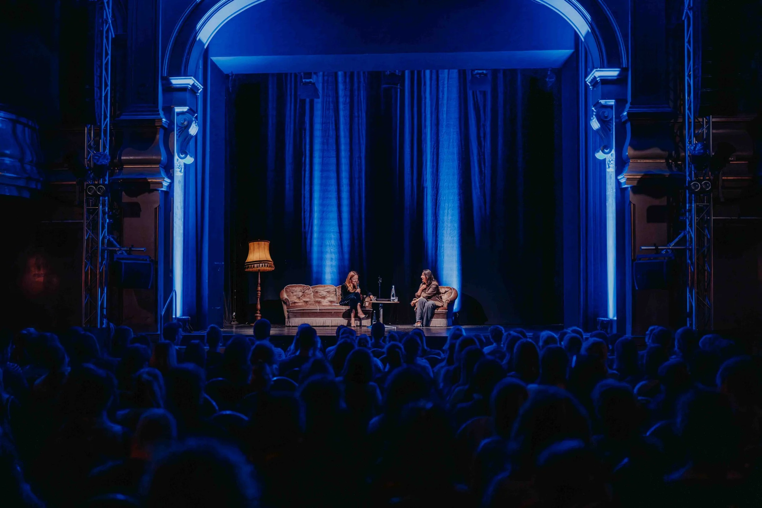 Theatersaal in blauem Licht mit vollem Publikum und Podiumsgästen auf der Bühne