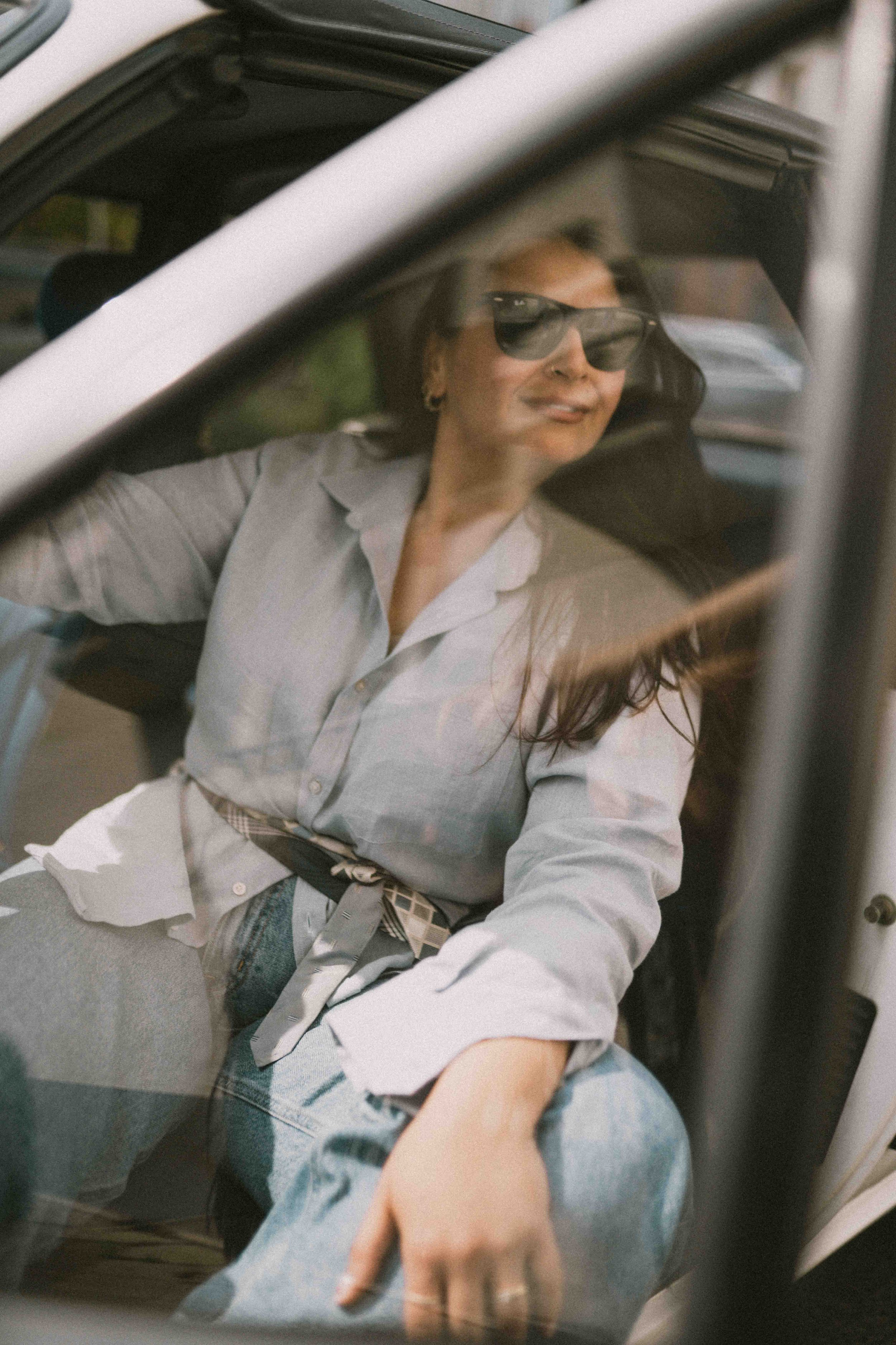 Person mit Sonnenbrille sitzt in einem Auto, trägt hellen Blazer
