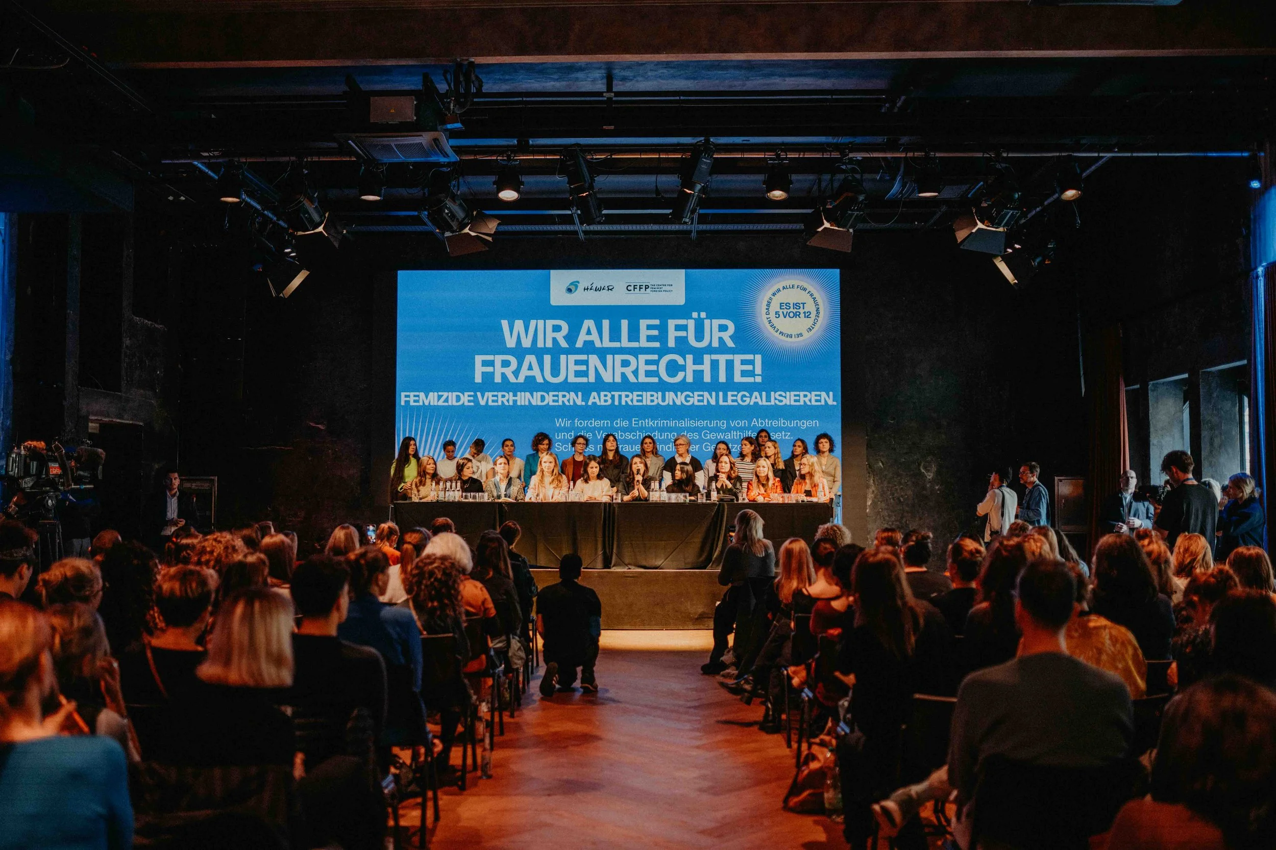 Veranstaltungsraum mit Publikum, im Hintergrund eine Leinwand mit der Aufschrift "Wir alle für Frauenrechte"