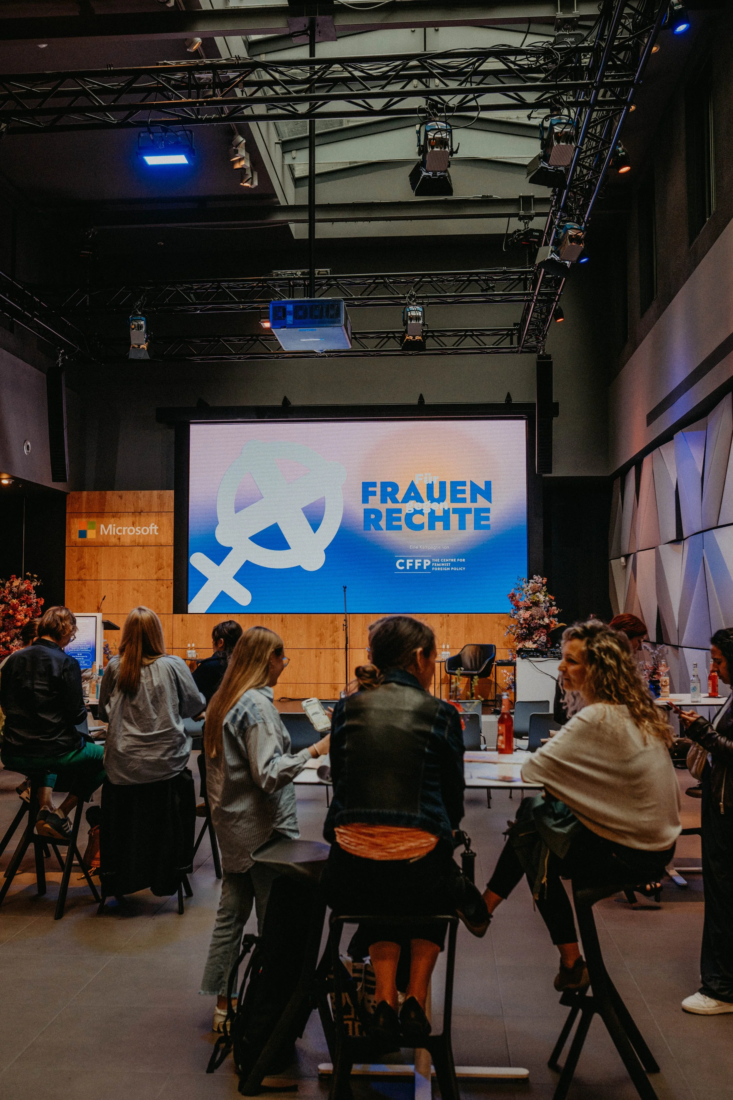 Publikum sitzt in einer Halle, im Hintergrund eine Leinwand mit dem Kampagnenlogo „Frauen Rechte"