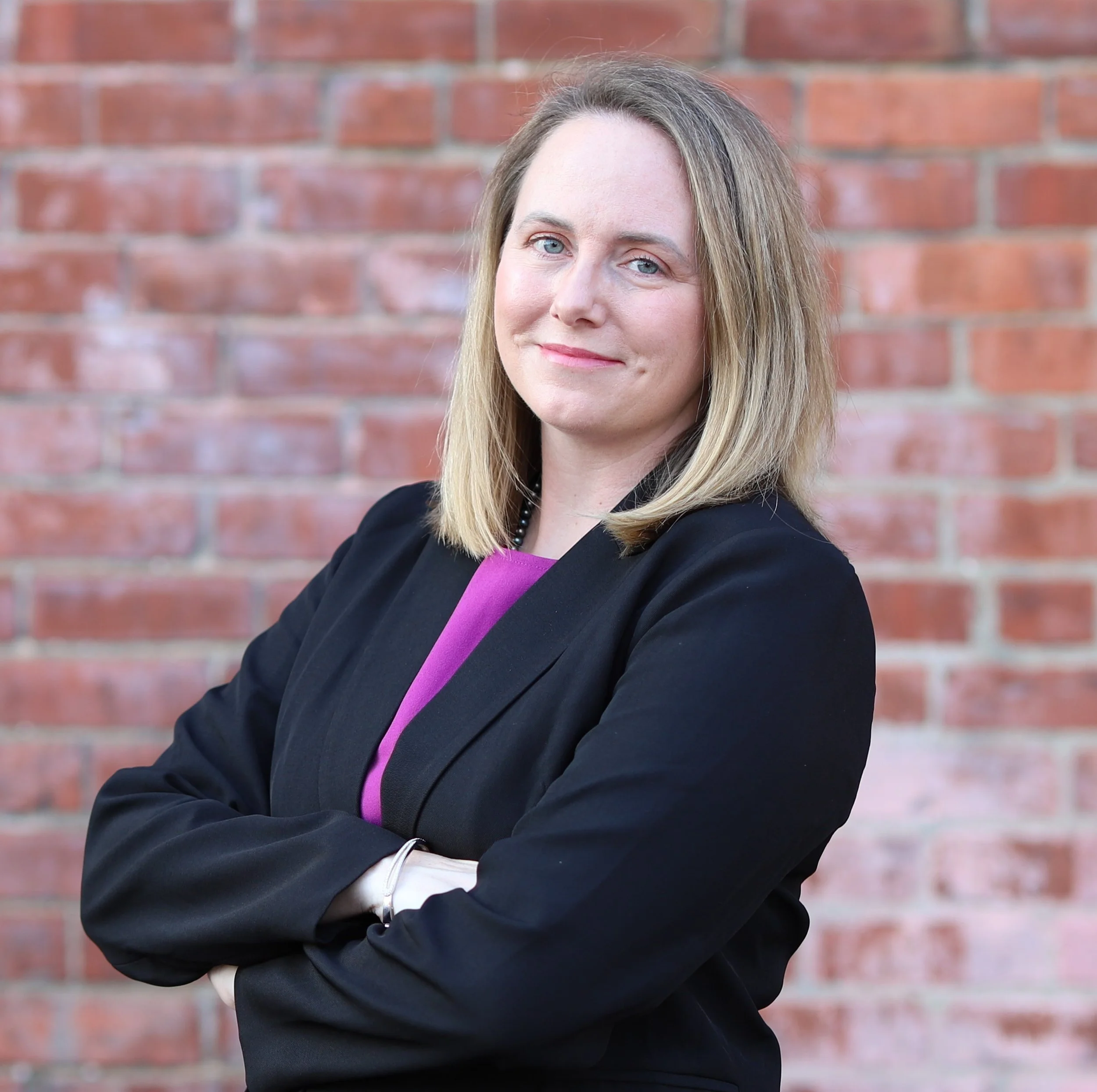 A woman with blonde hair and blue eyes is standing in front of a brick wall. She is wearing a black blazer, a purple top, and a beaded necklace. She has a slight smile and is looking into the camera.