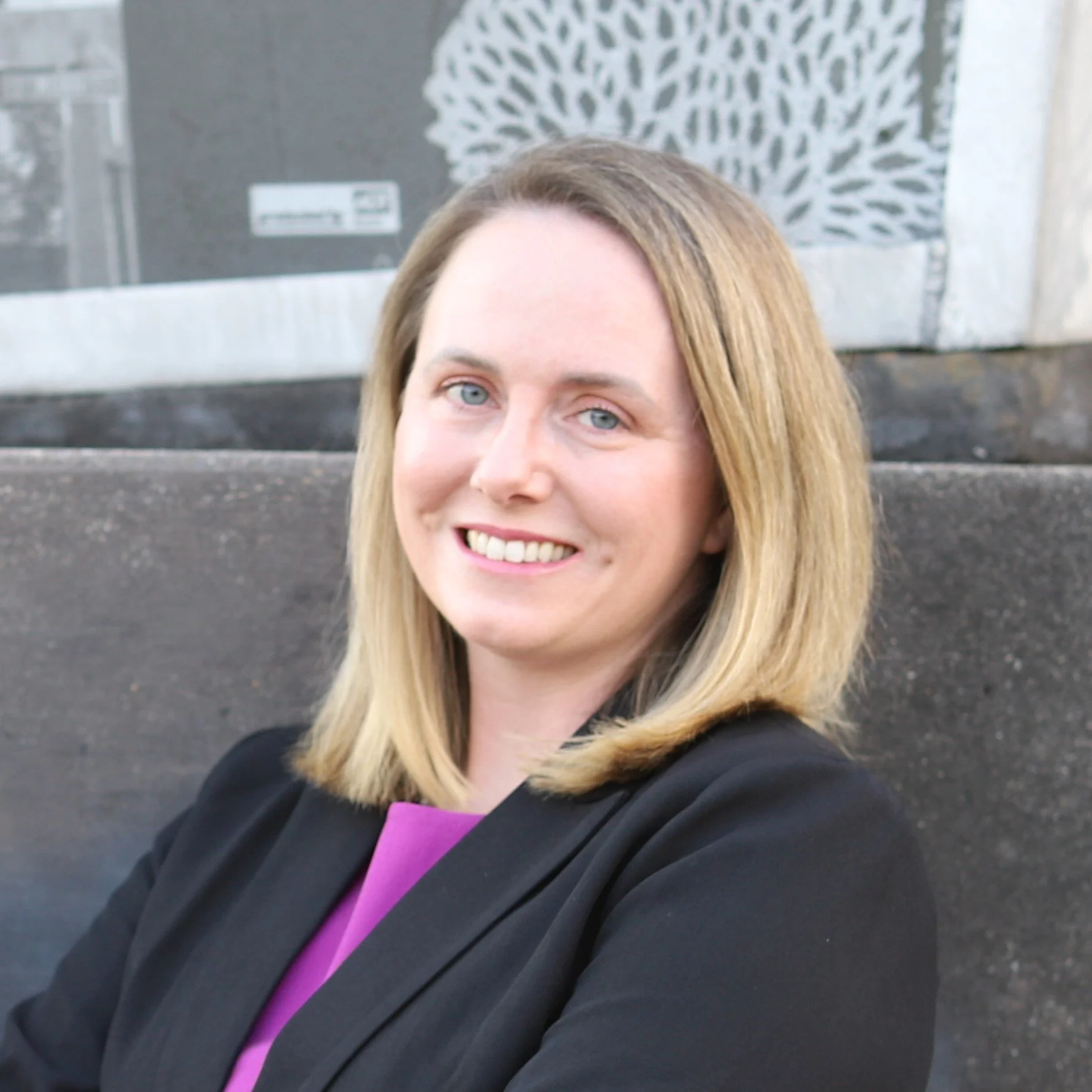 A woman with blonde hair and blue eyes smiling, wearing a black jacket with a purple shirt underneath, standing outside in front of concrete stairs and a wall with abstract white and gray artwork.