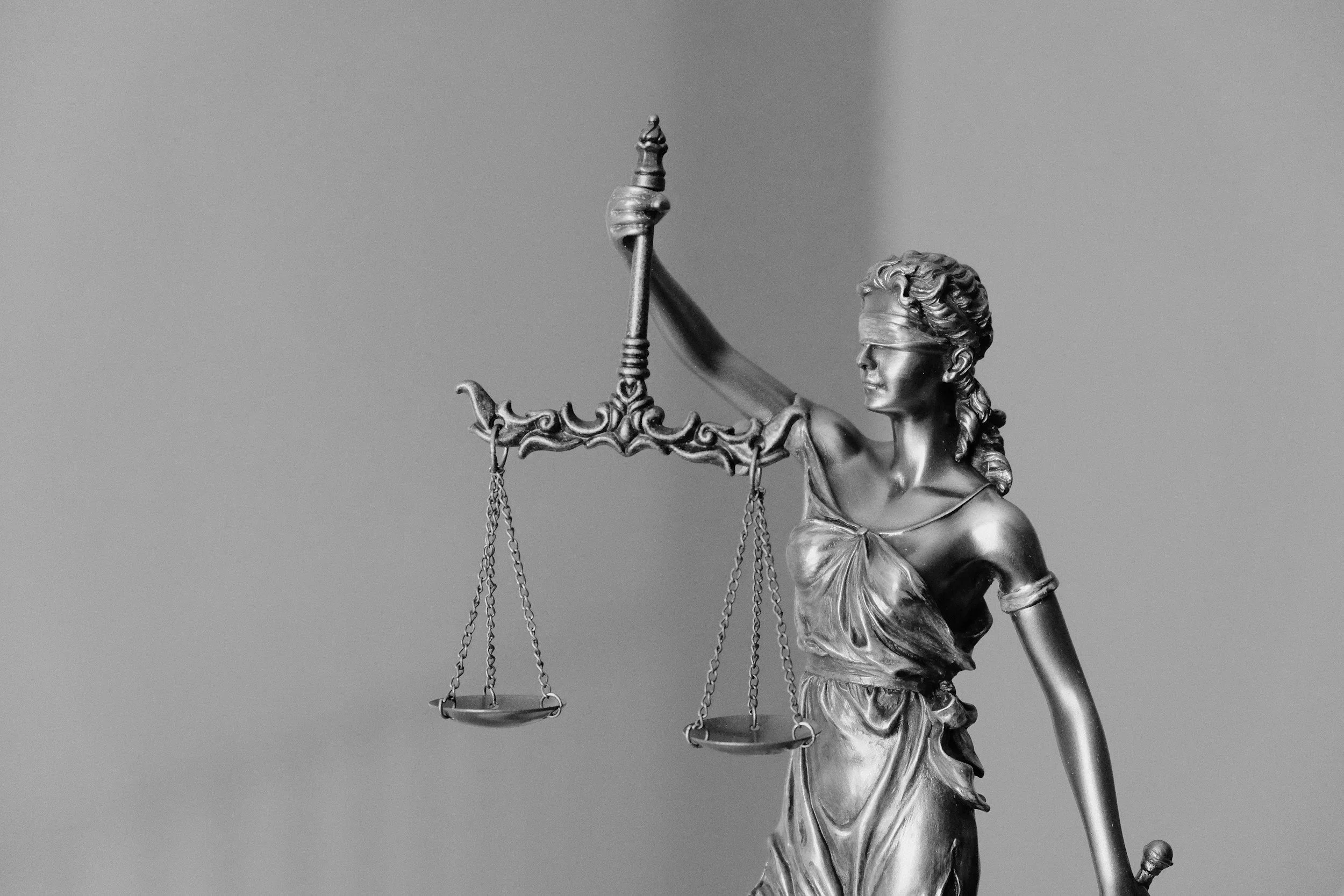 A metallic statue of Lady Justice with a blindfold, holding a set of scales in her left hand and a sword in her right hand, against a plain background.