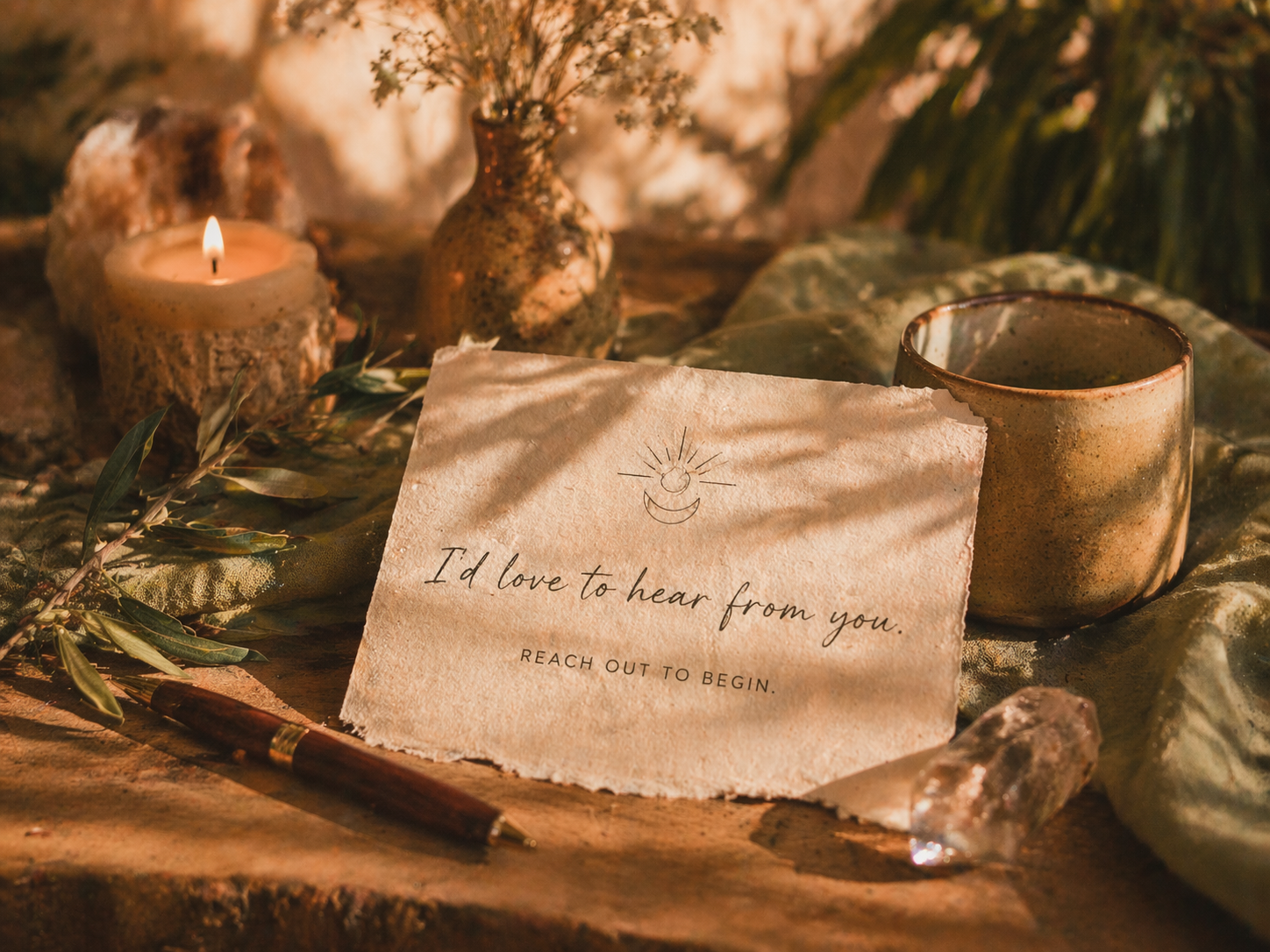 A handwritten note on textured paper that says, 'I'd love to hear from you. Reach out to begin.' The note has a sun and moon drawing at the top. Surrounding the note are a lit candle in a rustic holder, a ceramic cup, a small decorative jar, some foliage, a pen, and a crystal, all set on a fabric surface with a warm, ambient glow.