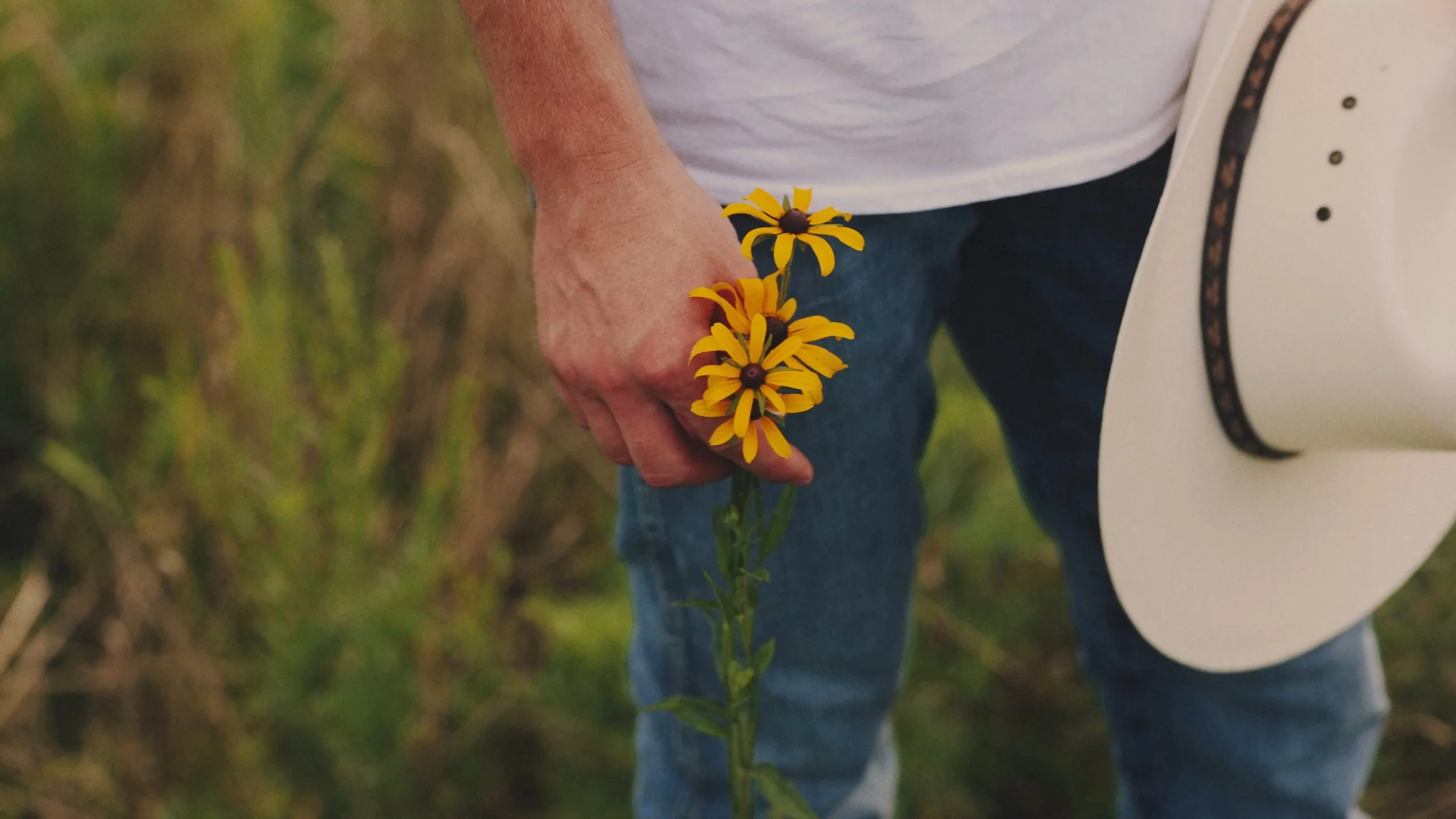 A man with a cowboy hat in one hand and a bunch of yellow wildflowers in the other hand.