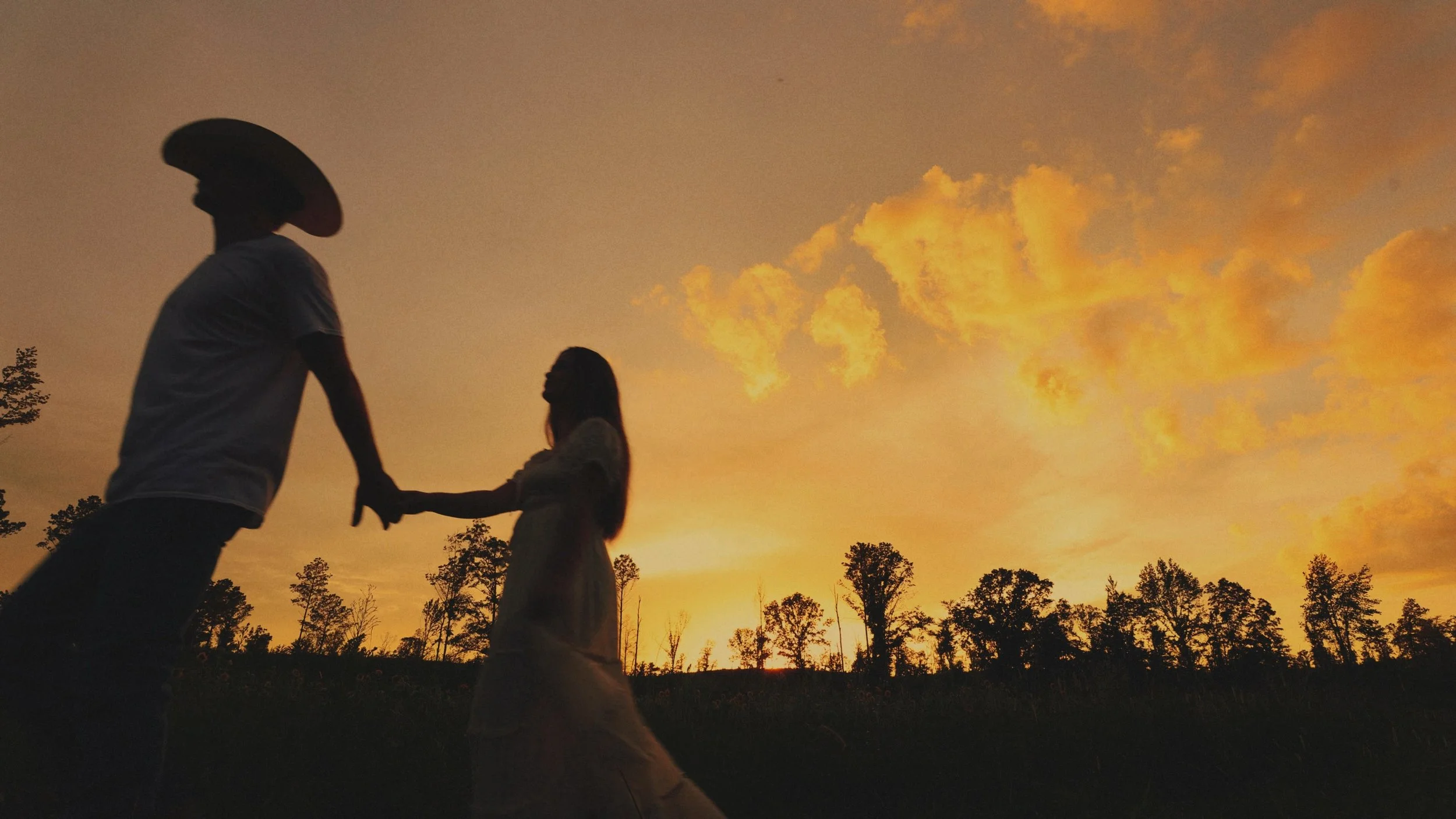 A silhouette of a bride and groom holding hands and walking with a beautiful yellow and orange sunset in the sky. 