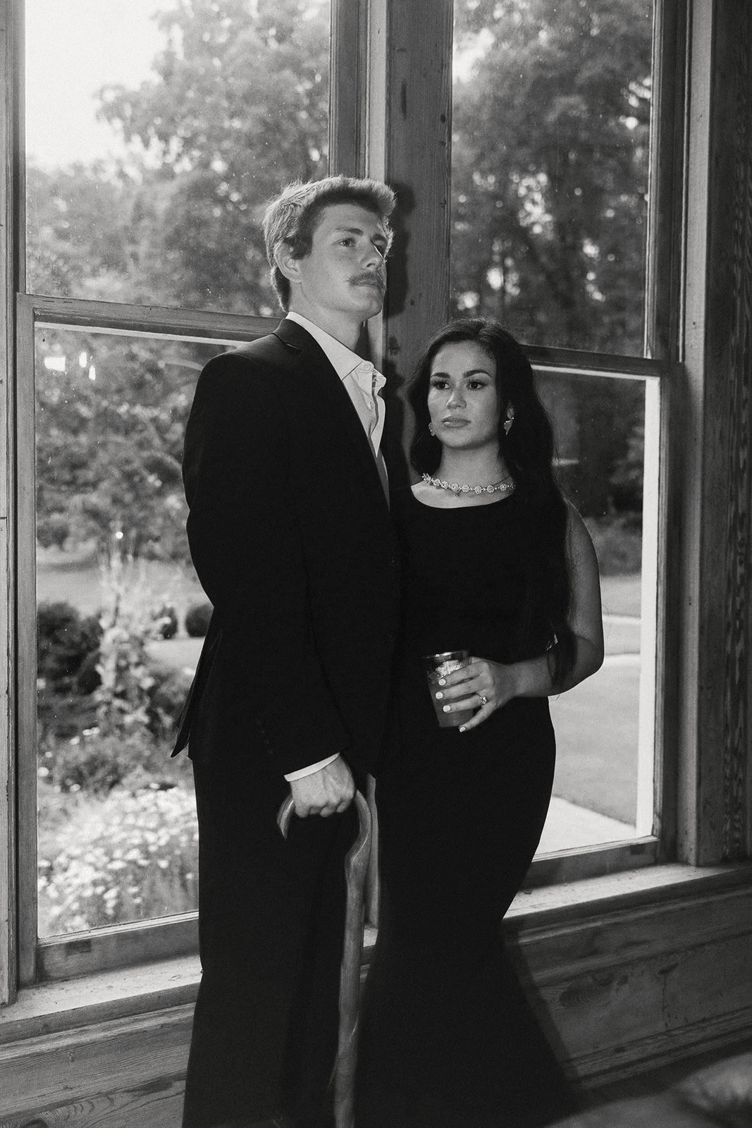 Black and white image of a couple dressed in black tie attire posing seriously in front of a window.