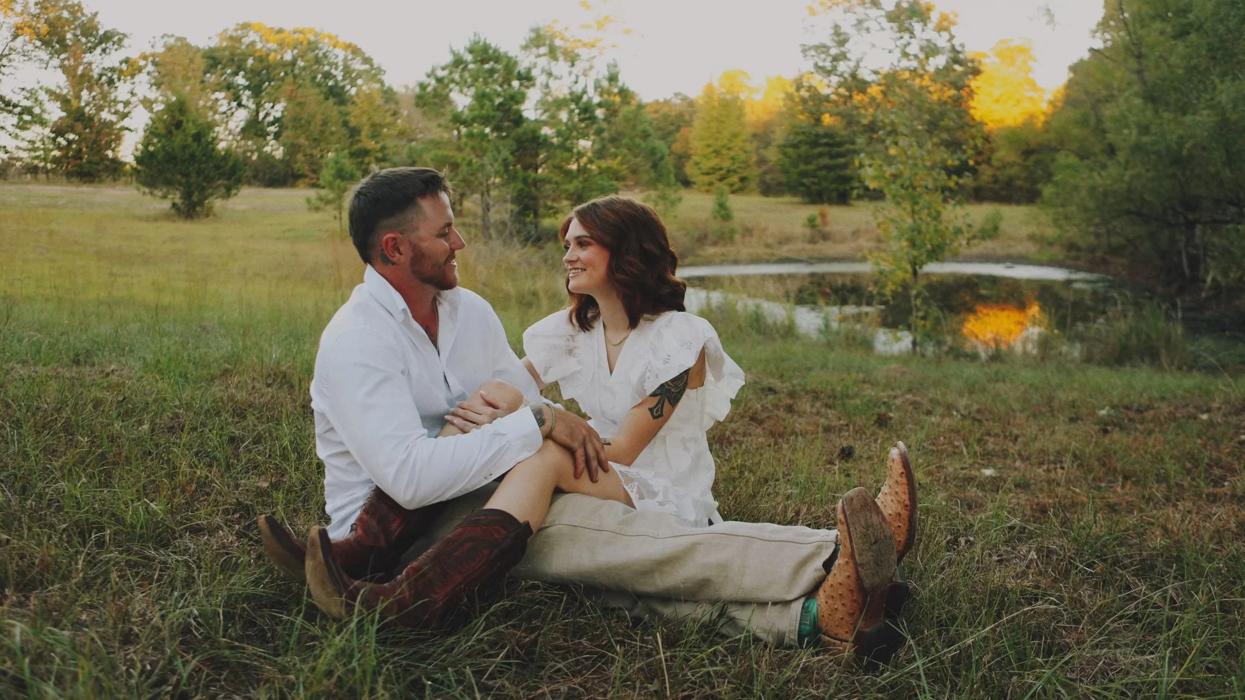 A couple sitting on the ground looking at each other and smiling in front of a pond in Mississippi.