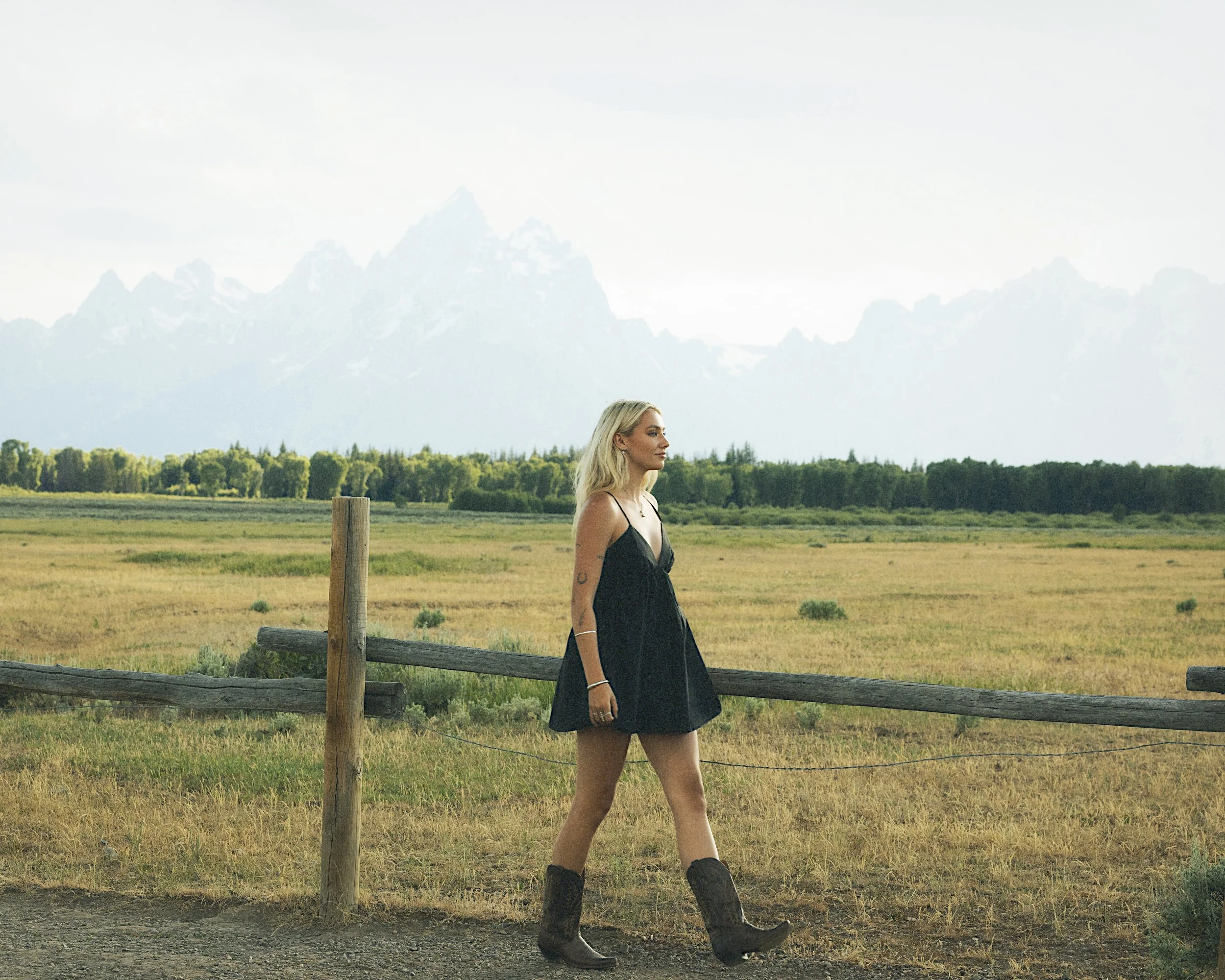 A woman in a black dress and cowboy boots walking in front of a pasture and mountain range.