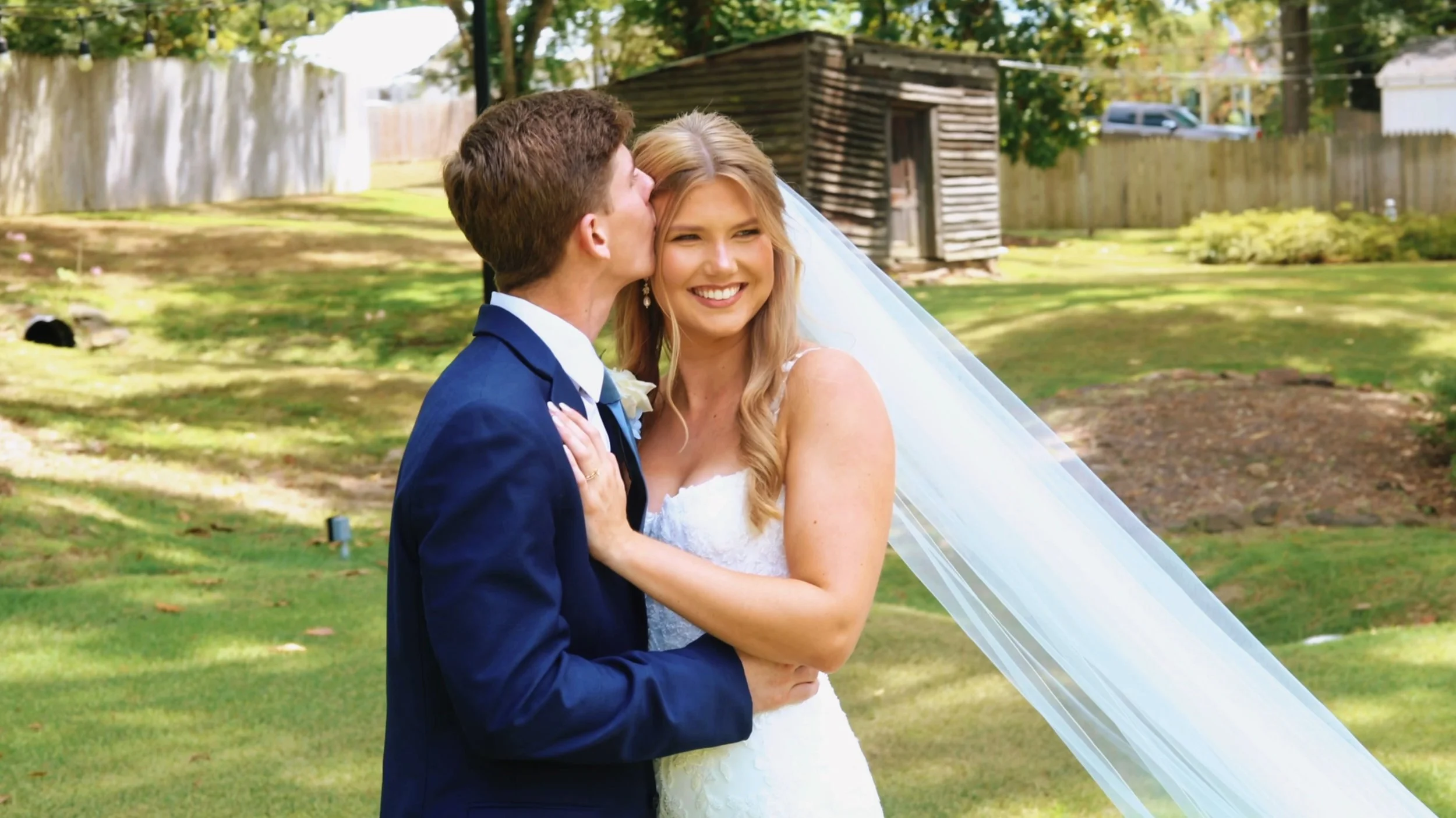 A groom is giving his bride a kiss on the cheek while she smiles out into the distance.