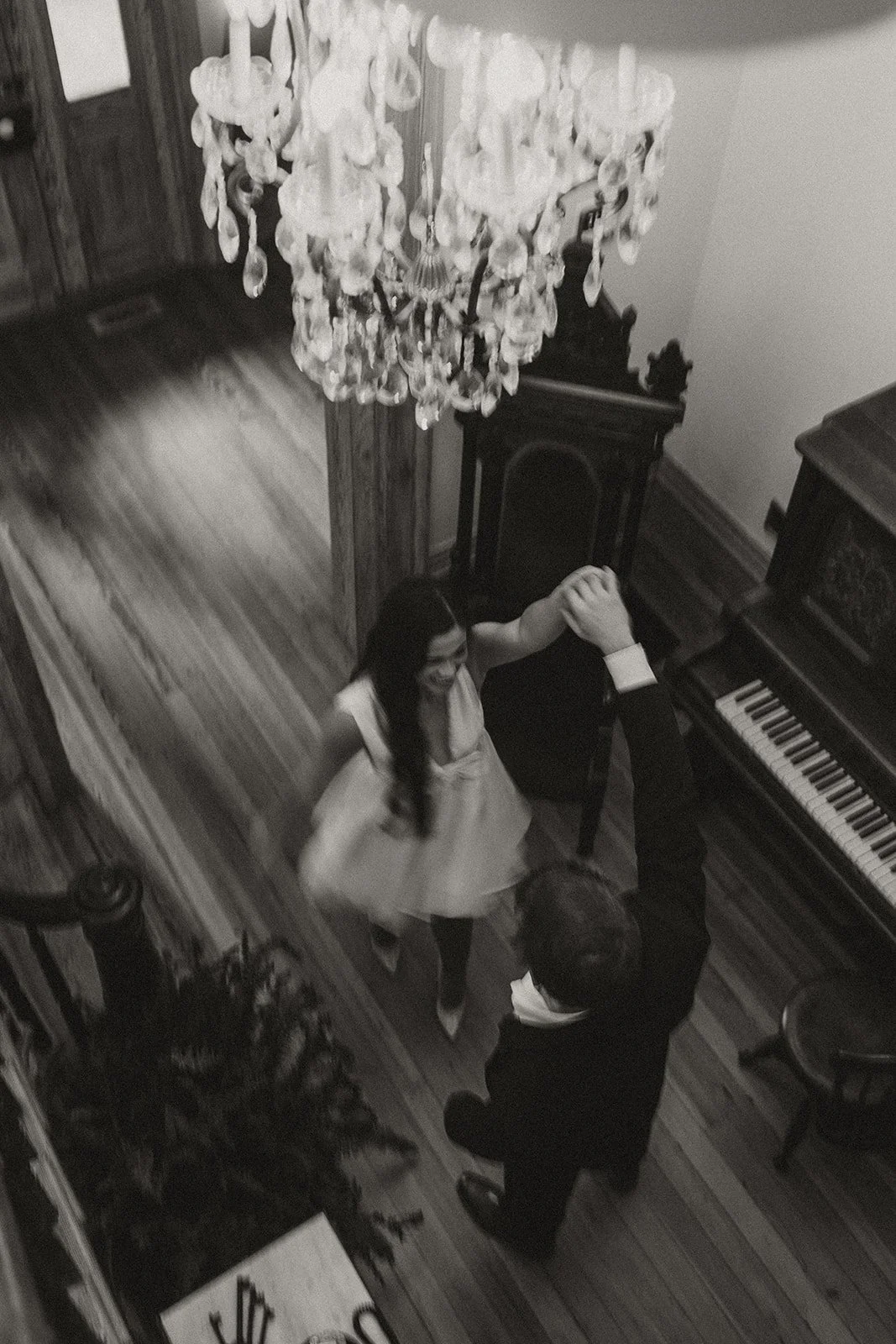 Black and white image of a couple dancing underneath a chandelier, beside a grand piano.