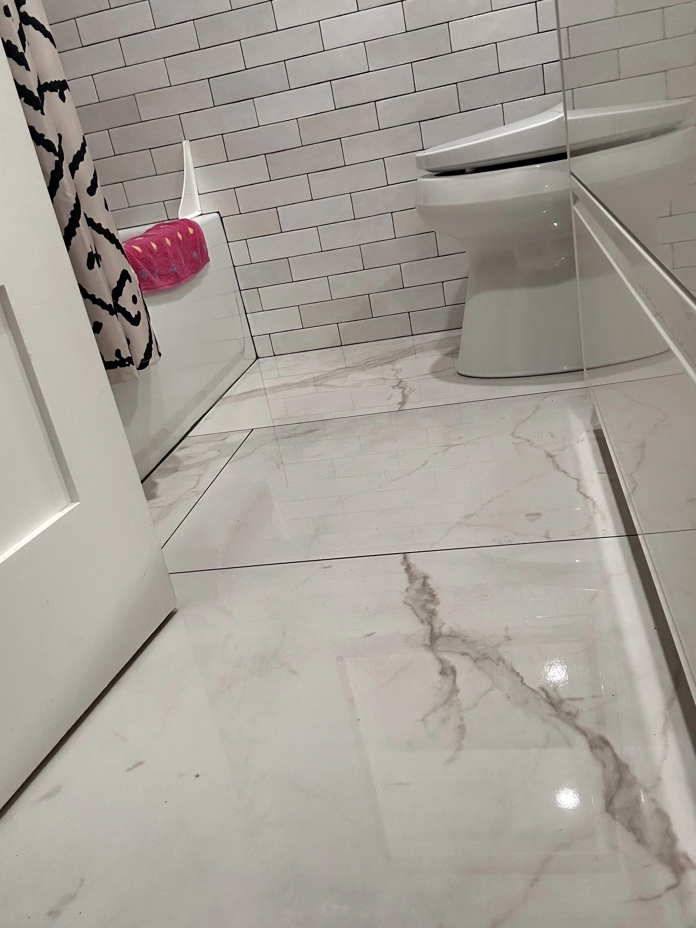 A bathroom with a white toilet, marble floor, and white tile walls. A shower with a pink soap holder and a towel hanging on the shower door.