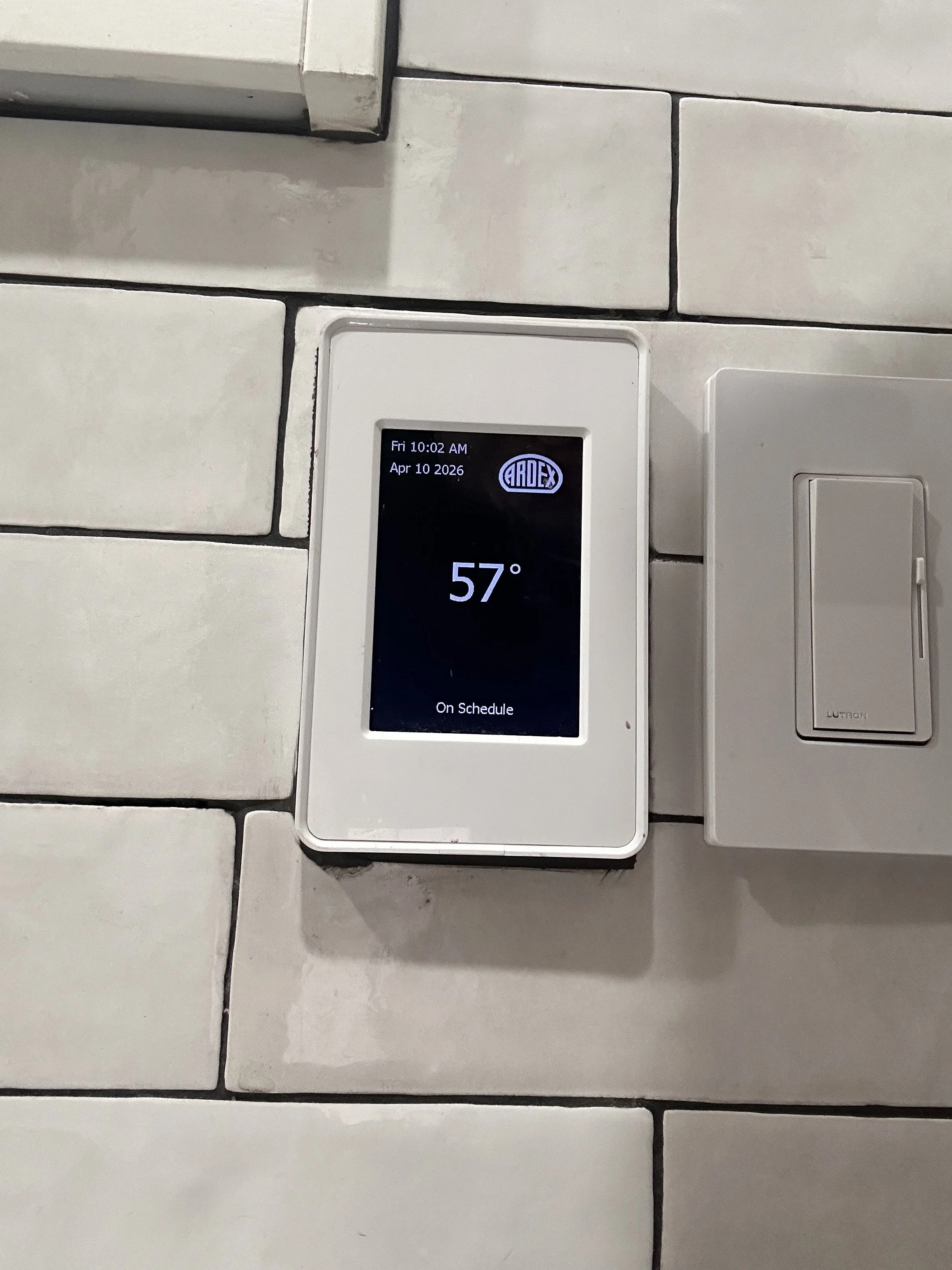 Digital thermostat mounted on a white tiled wall displaying a temperature of 57°F, labeled 'On Schedule,' with the date and time showing 10:02 AM on April 10, 2026.