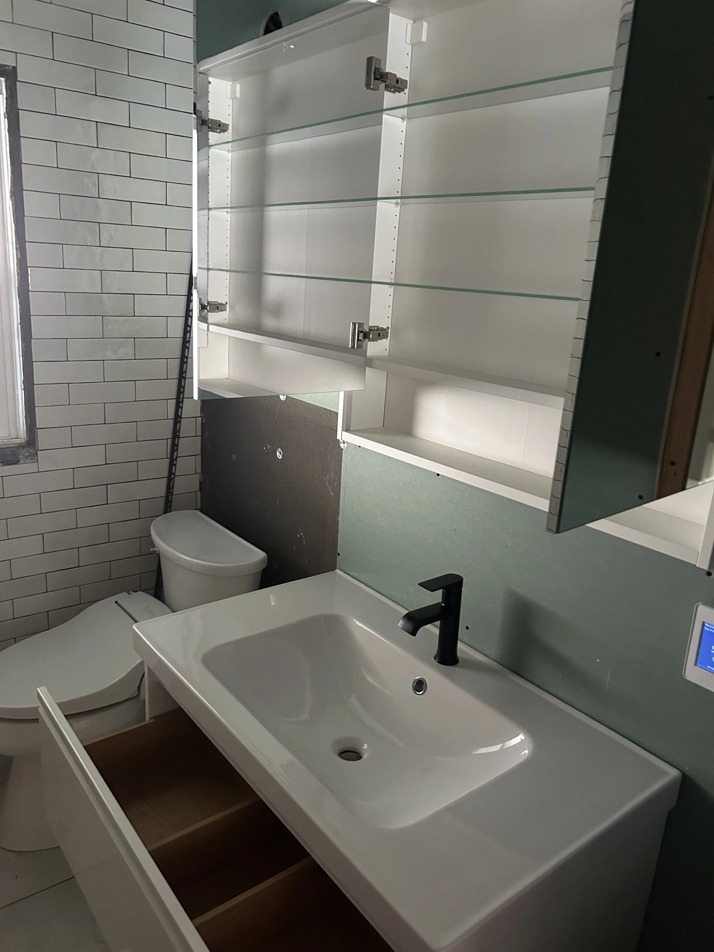 Bathroom with a white sink, black faucet, toilet, and a mounted white cabinet with glass shelves, beside a window with white tile wall.
