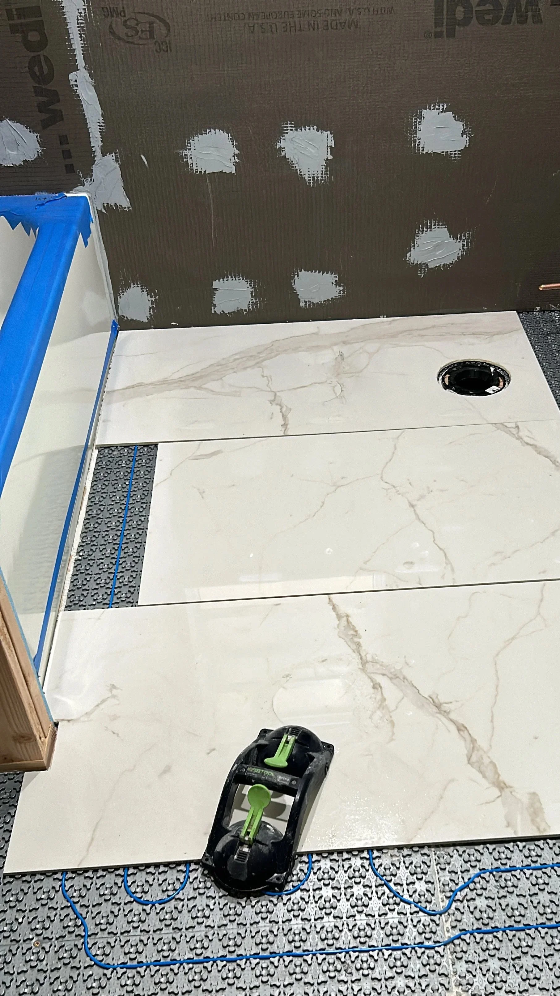 Installing marble tiles on a floor with a black drain hole. A powder tool is placed on the tiles. Blue electrical wires run underneath the tiles on a textured black surface.