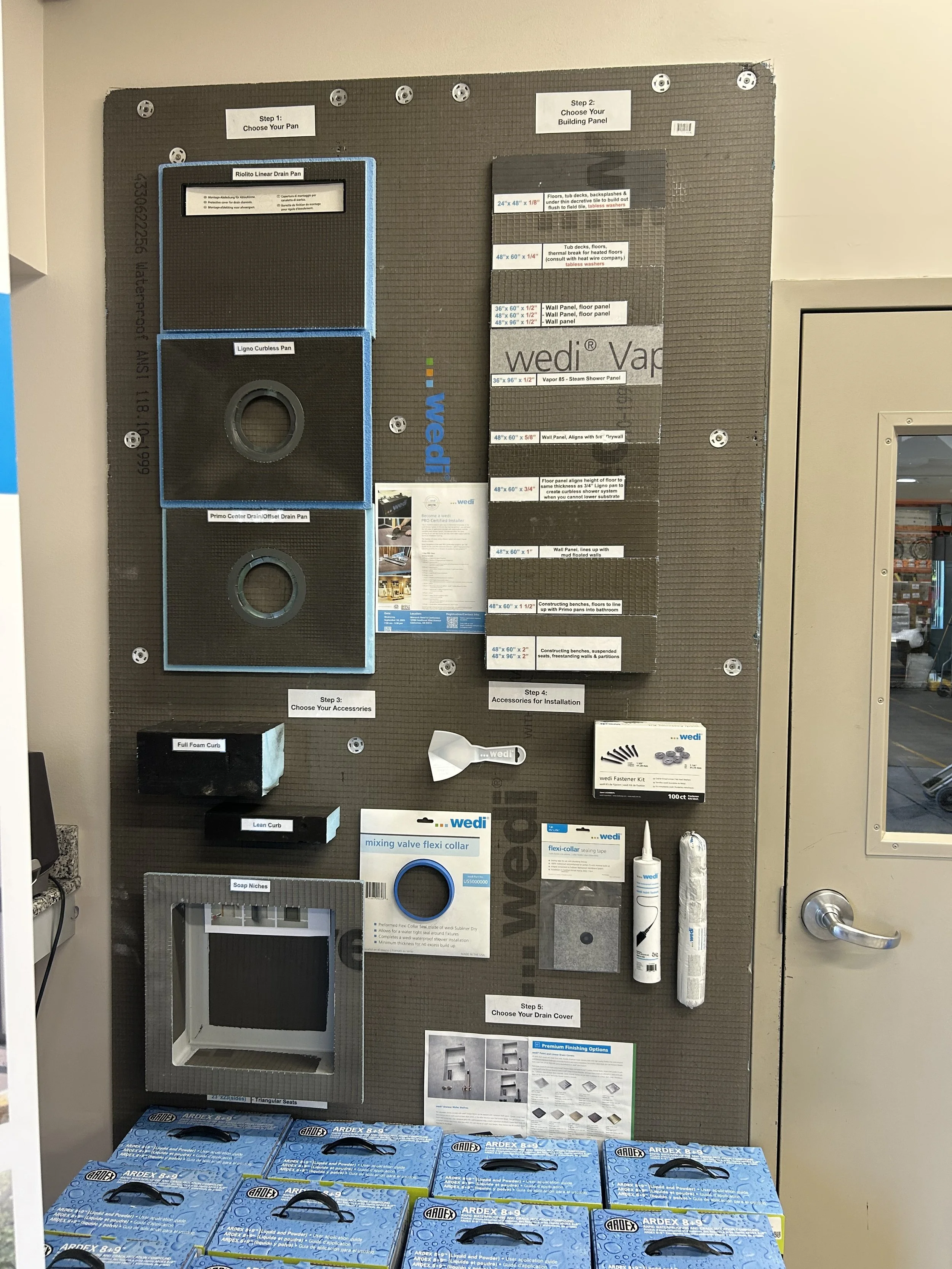 Display board with steps and supplies for building a shower or plumbing project, including drain pans, wall panels, accessories, and installation tools. Bowls of boxes of caulk or sealant at the bottom.
