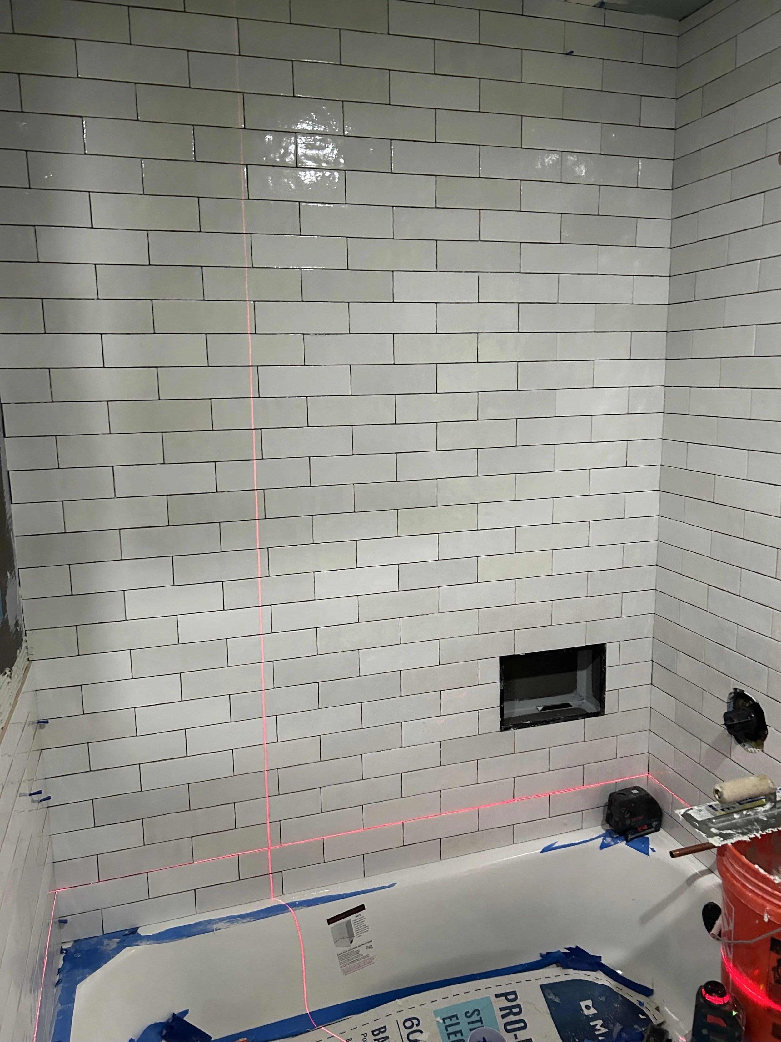 Bathroom under construction with white subway tile wall, red laser level lines, and an opening for a plumbing fixture on the wall.