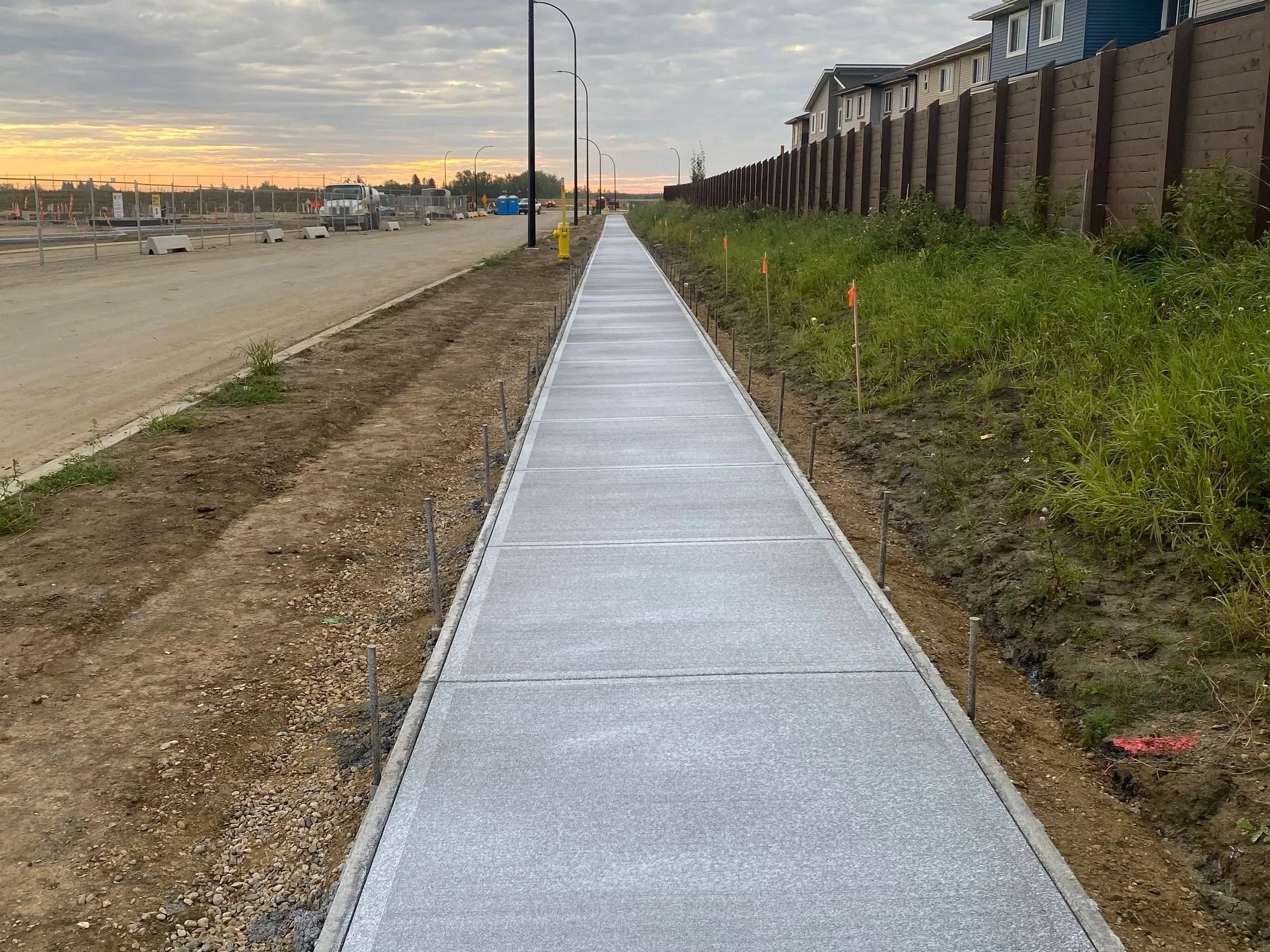 A newly constructed concrete sidewalk runs through a developing area with grass and dirt on either side, residential buildings on the right, and a dirt lot with construction equipment on the left, during sunset.