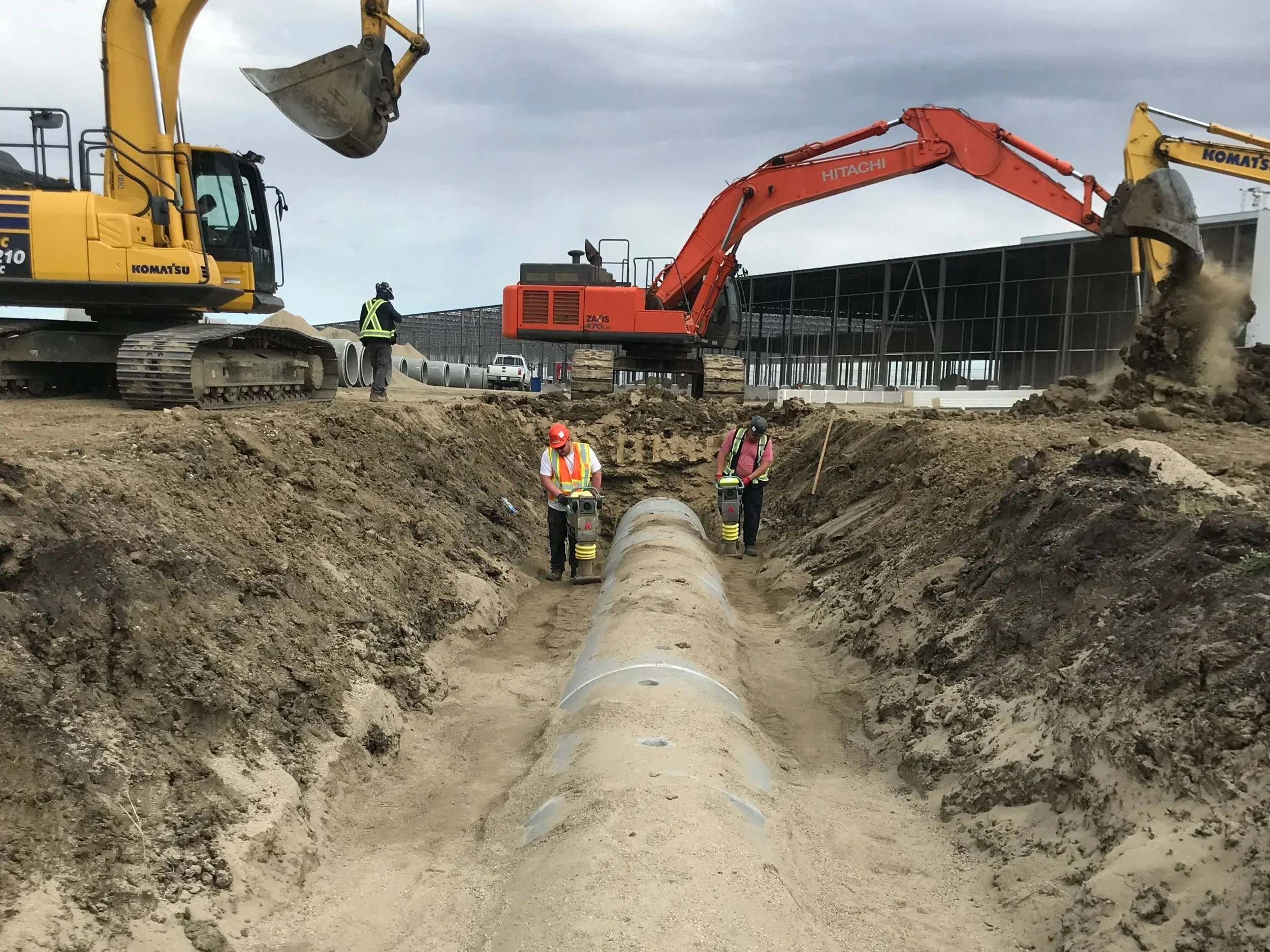 Construction site with large excavator and front loader working on underground pipeline installation. Two workers wearing safety gear inspecting the pipe in the trench.