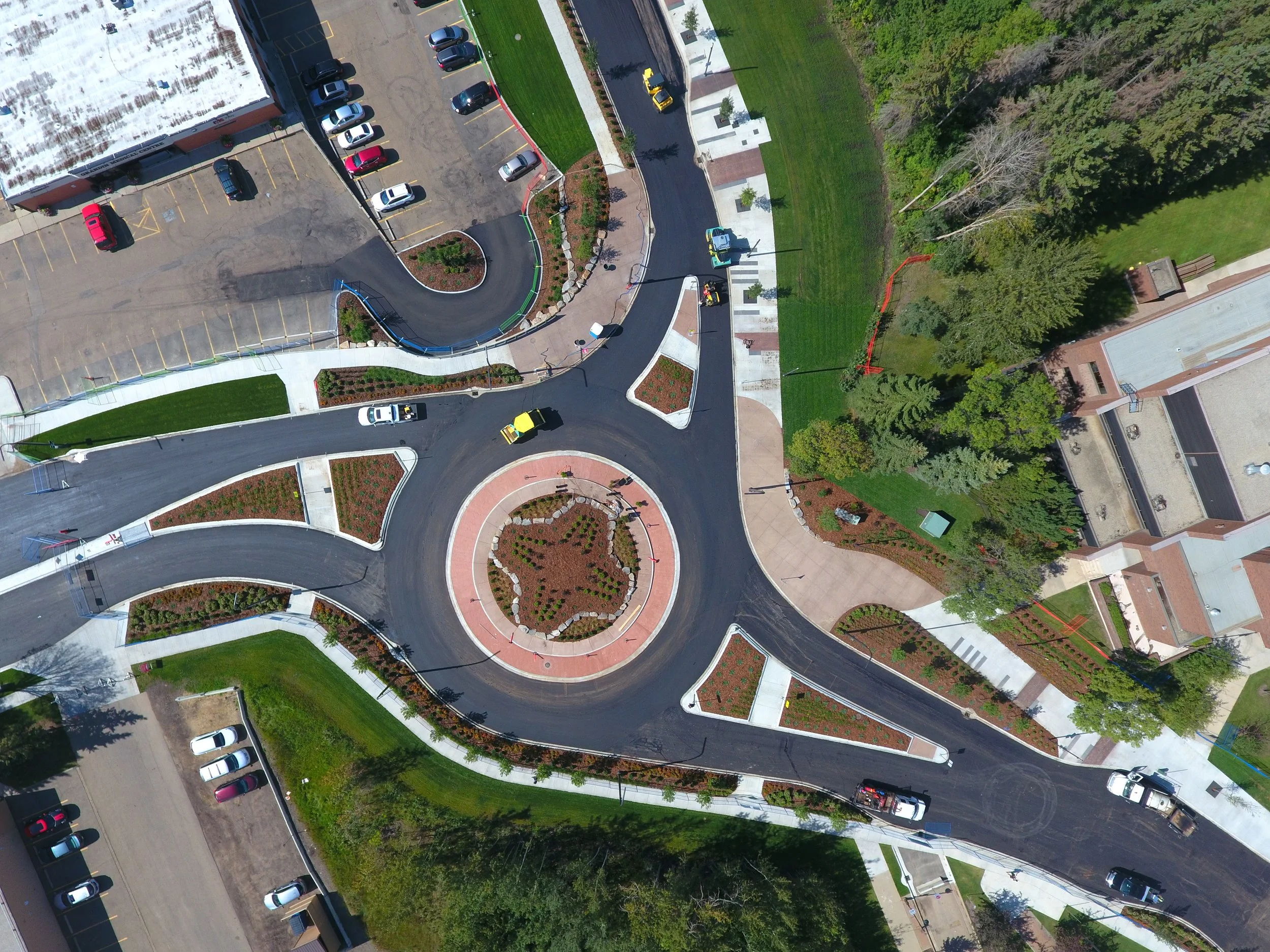 An aerial view of a new roundabout with landscaped islands, sidewalks, grass, trees, and parked cars.