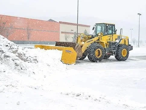 Yellow snowplow clearing snow from a parking lot with a red brick building in the background.