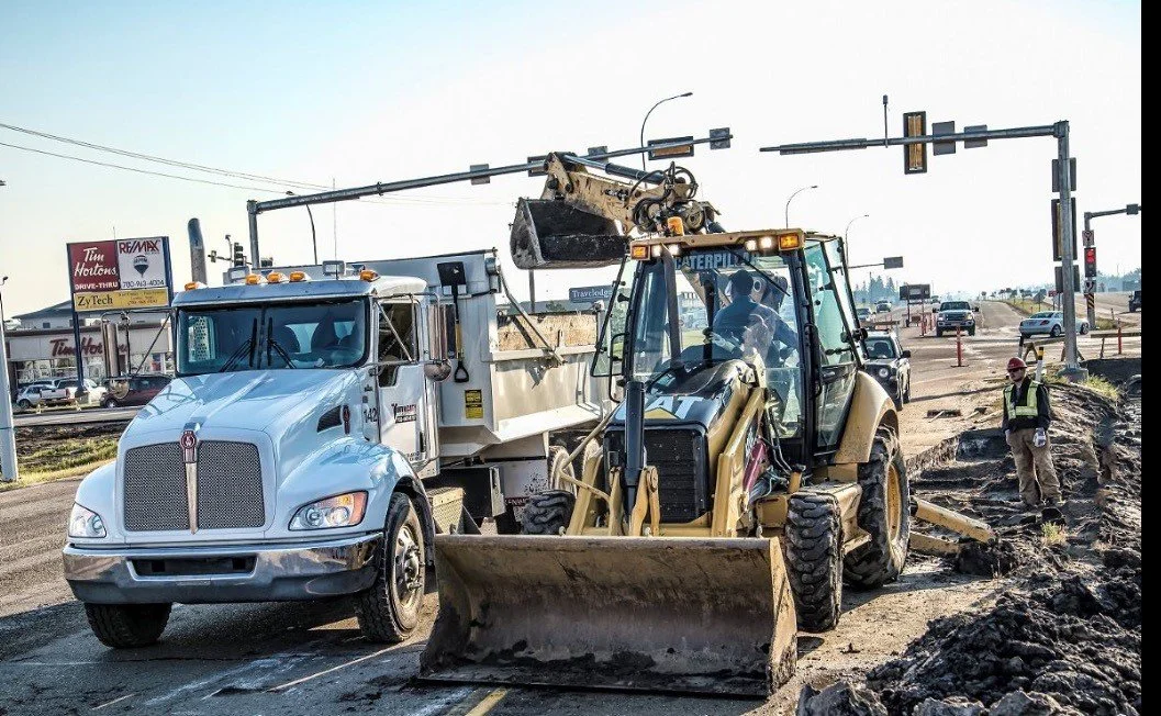 Construction vehicles working on a road, with a bulldozer and a dump truck, and a construction worker nearby.