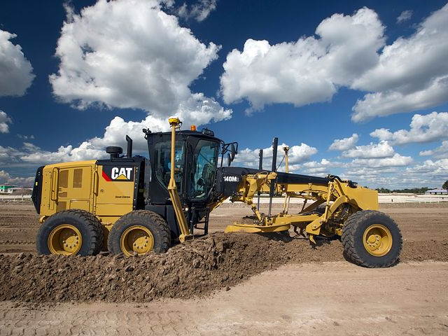 A yellow Caterpillar motor grader working on a dirt road against a cloudy sky.
