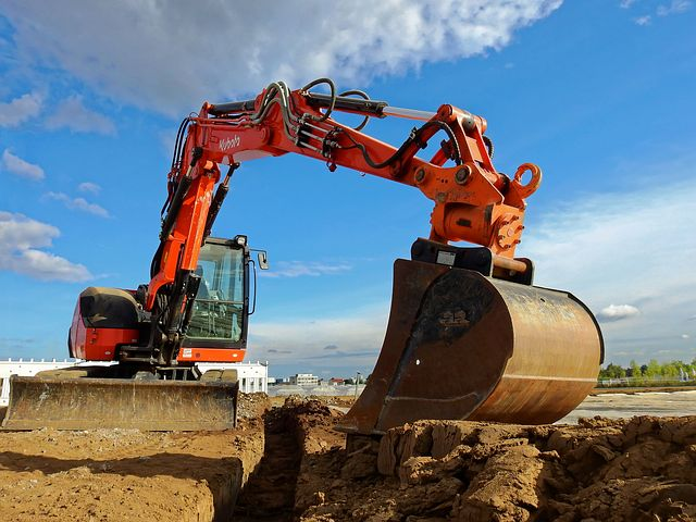 An orange excavator digging into the dirt on a construction site under a partly cloudy sky.