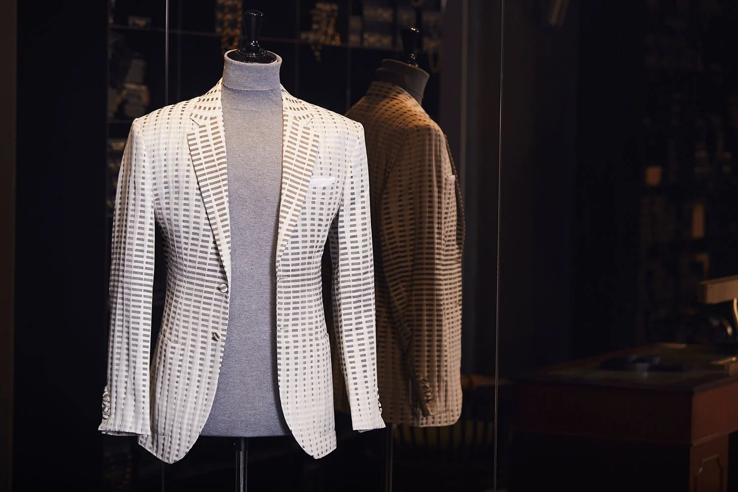 Mannequin dressed in a white and black checkered blazer over a gray turtleneck, displayed in a glass case, with a second mannequin dressed in a similar checkered blazer in the background.