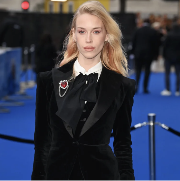A woman with blonde hair and light makeup dressed in a black velvet blazer with a brooch, standing on a blue carpet at an event.