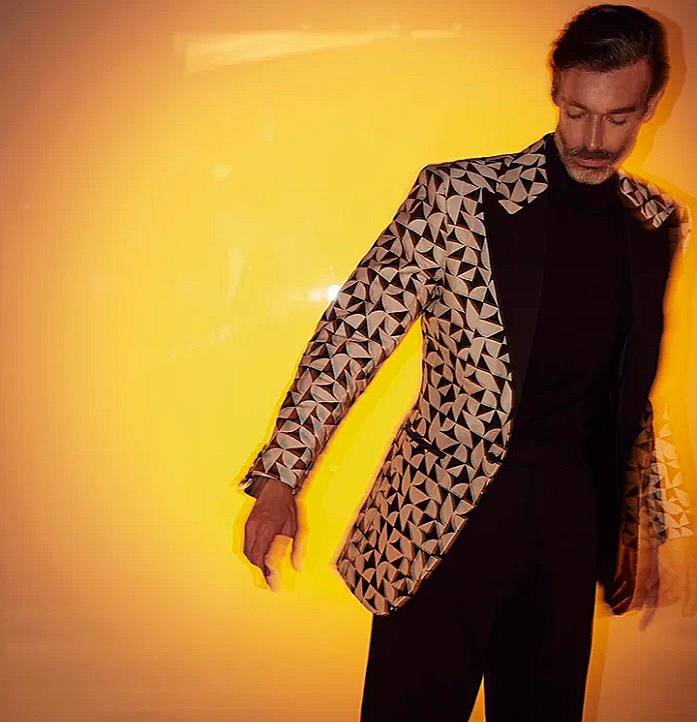 A man wearing a black turtleneck and a geometric patterned blazer standing against a yellow background.
