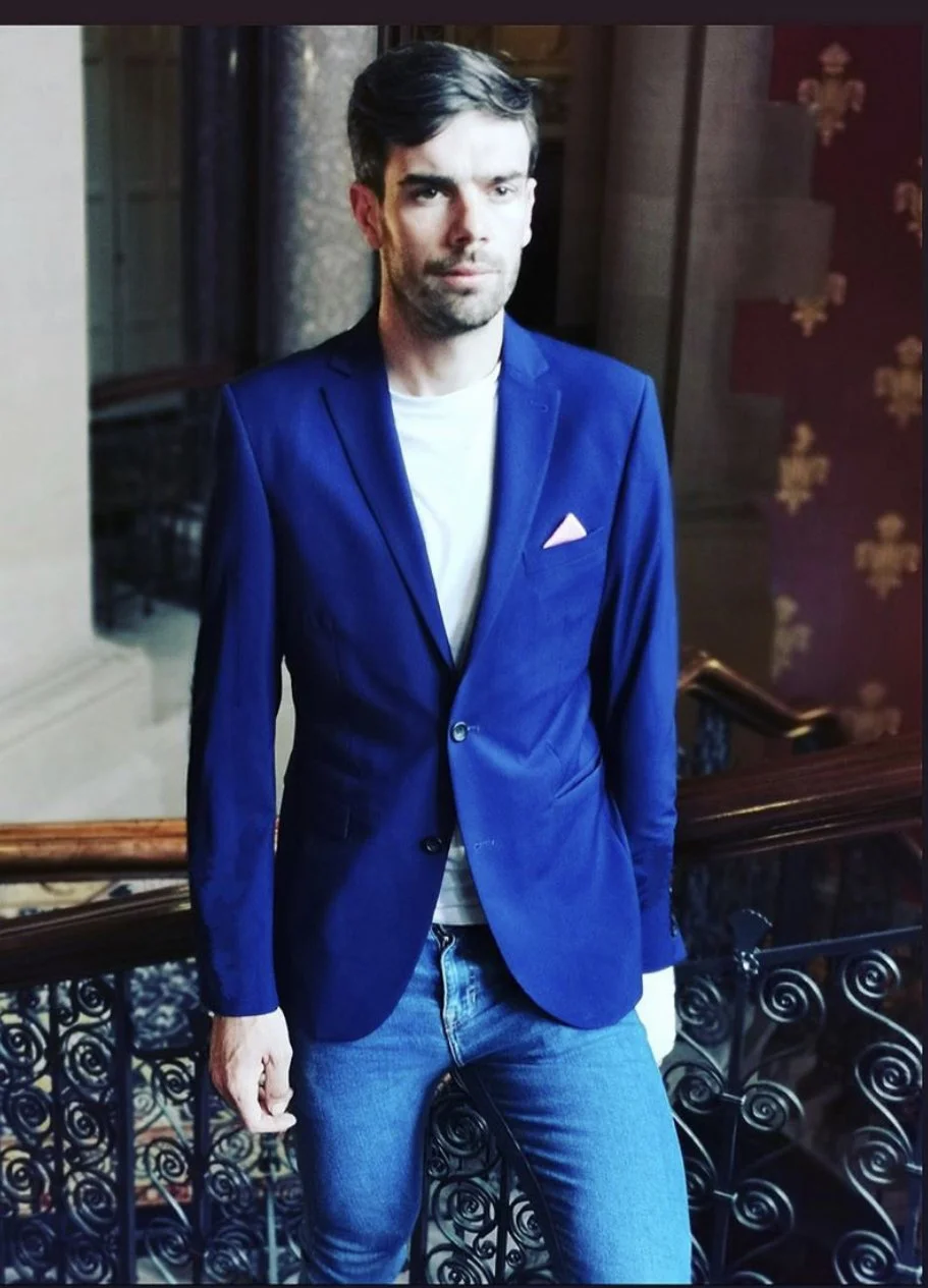 A man with dark hair and a beard, wearing a blue blazer, white shirt, and blue jeans, standing indoors near a staircase with a decorative railing and patterned wallpaper in the background.