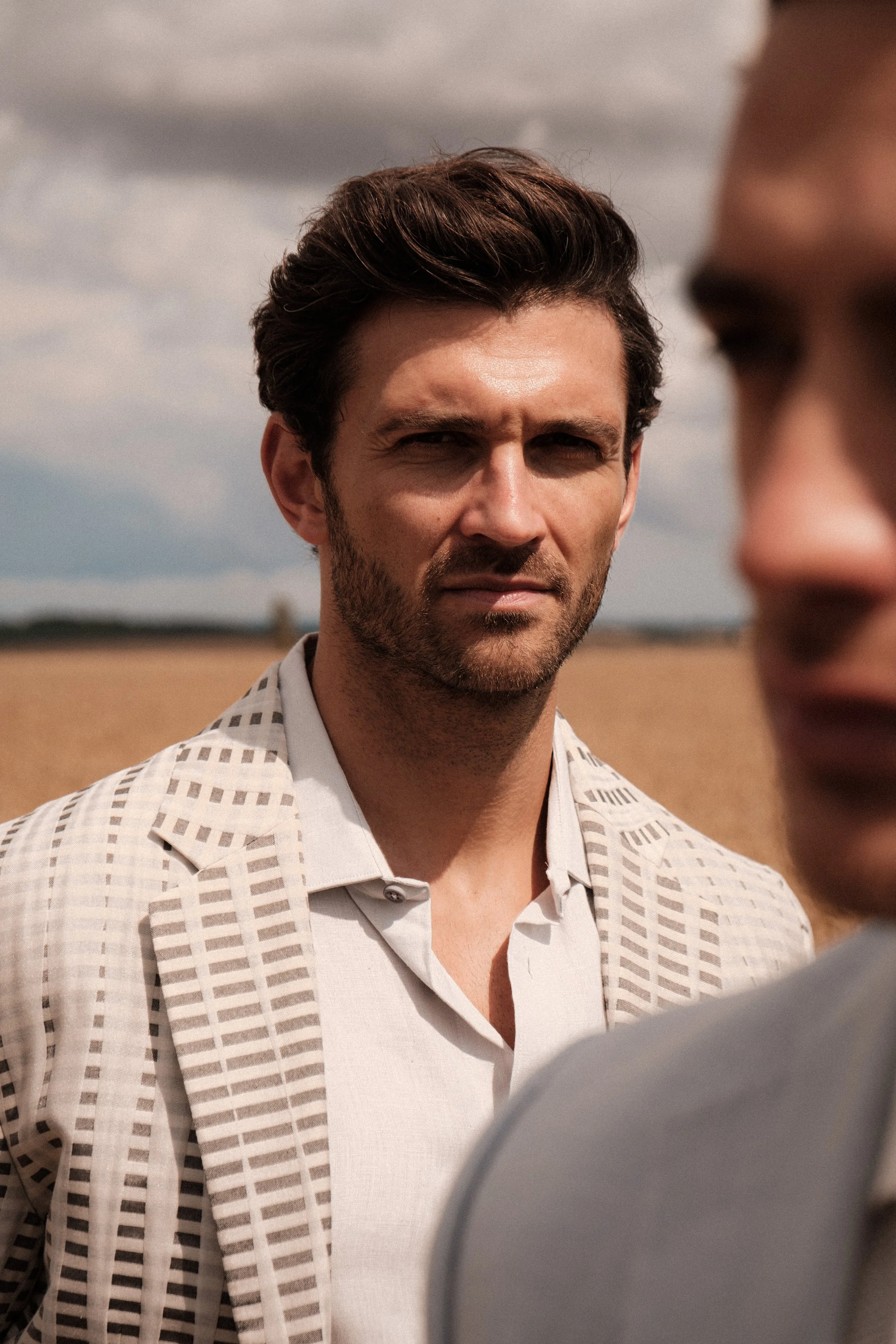 A man with dark brown hair and a beard, wearing a checkered blazer over a white shirt, looking serious in an outdoor setting with a cloudy sky.