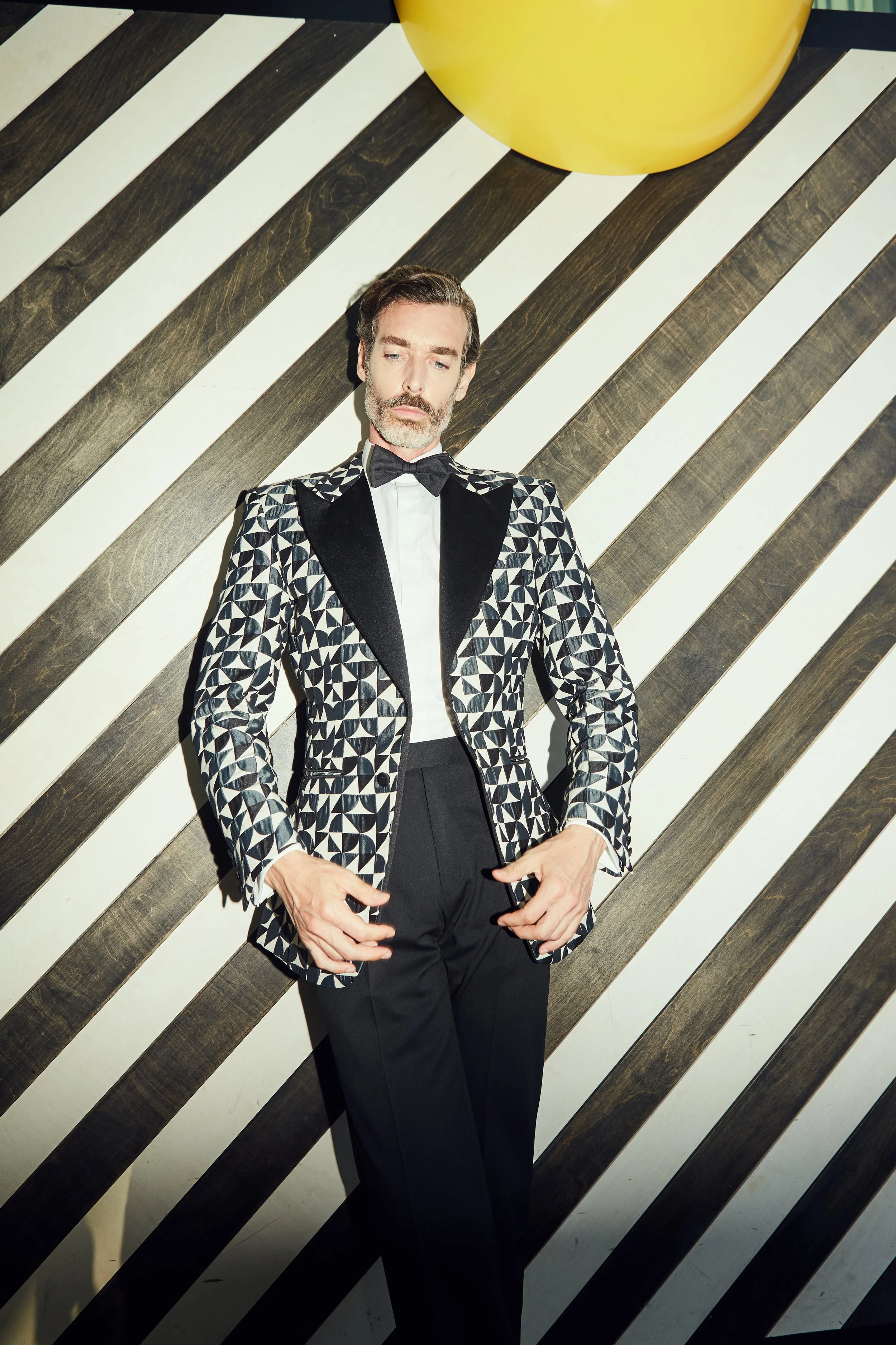 A man in a black and white geometric patterned tuxedo with a black bow tie, standing against a black and white striped wall with a large yellow circle above.