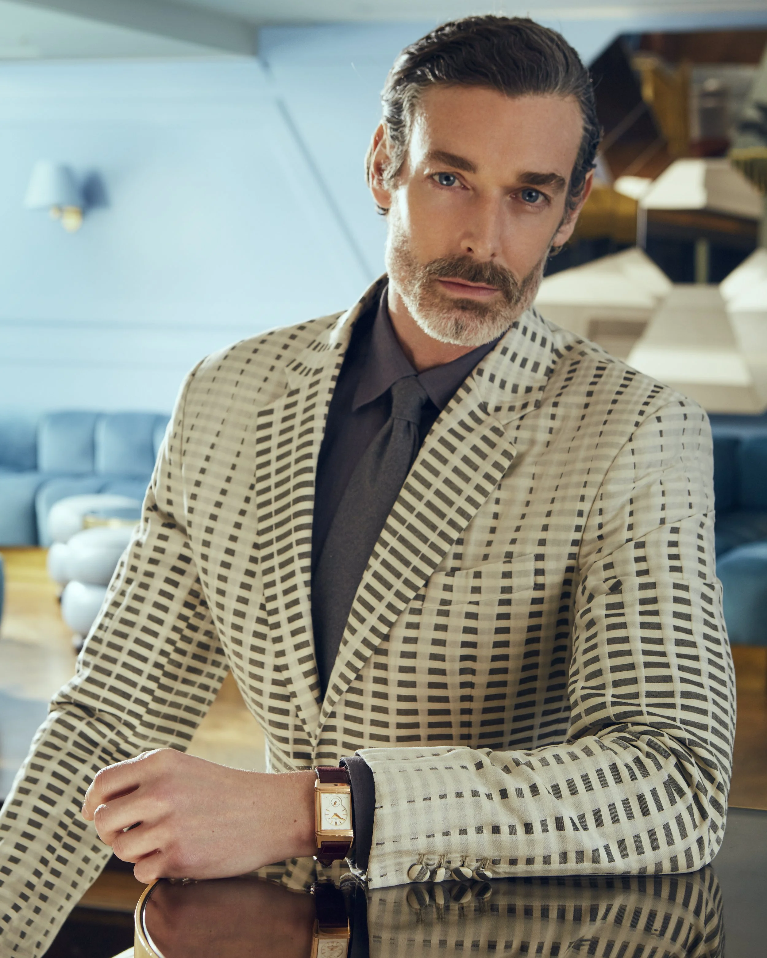 Close-up of a man wearing a checkered blazer and a dark shirt, with a watch on his wrist, in a modern, well-lit interior. The background features blue walls and contemporary furniture.