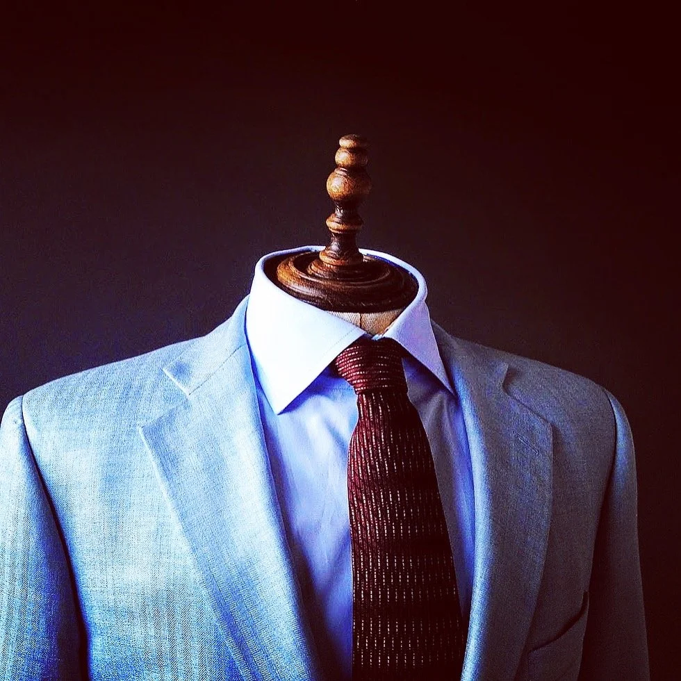 A mannequin dressed in a light gray suit, white shirt, and dark red tie, set against a dark background.