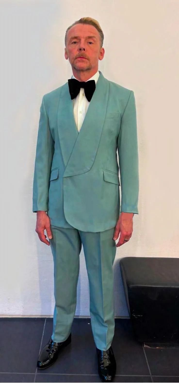 A man standing against a plain white wall, wearing a light blue suit with a black bow tie, white shirt, and black shiny dress shoes.