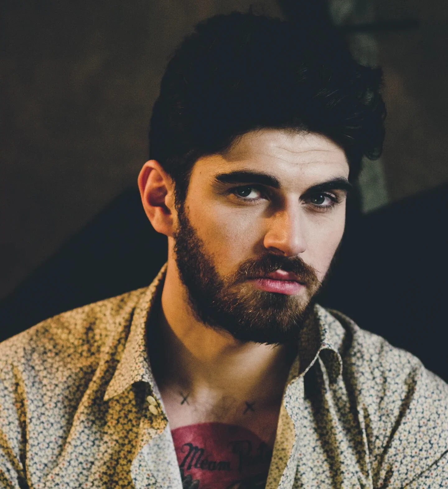 A close-up portrait of a man with dark hair, blue eyes, and a beard, wearing a patterned shirt with a red shirt underneath, looking directly at the camera in a dimly lit setting.