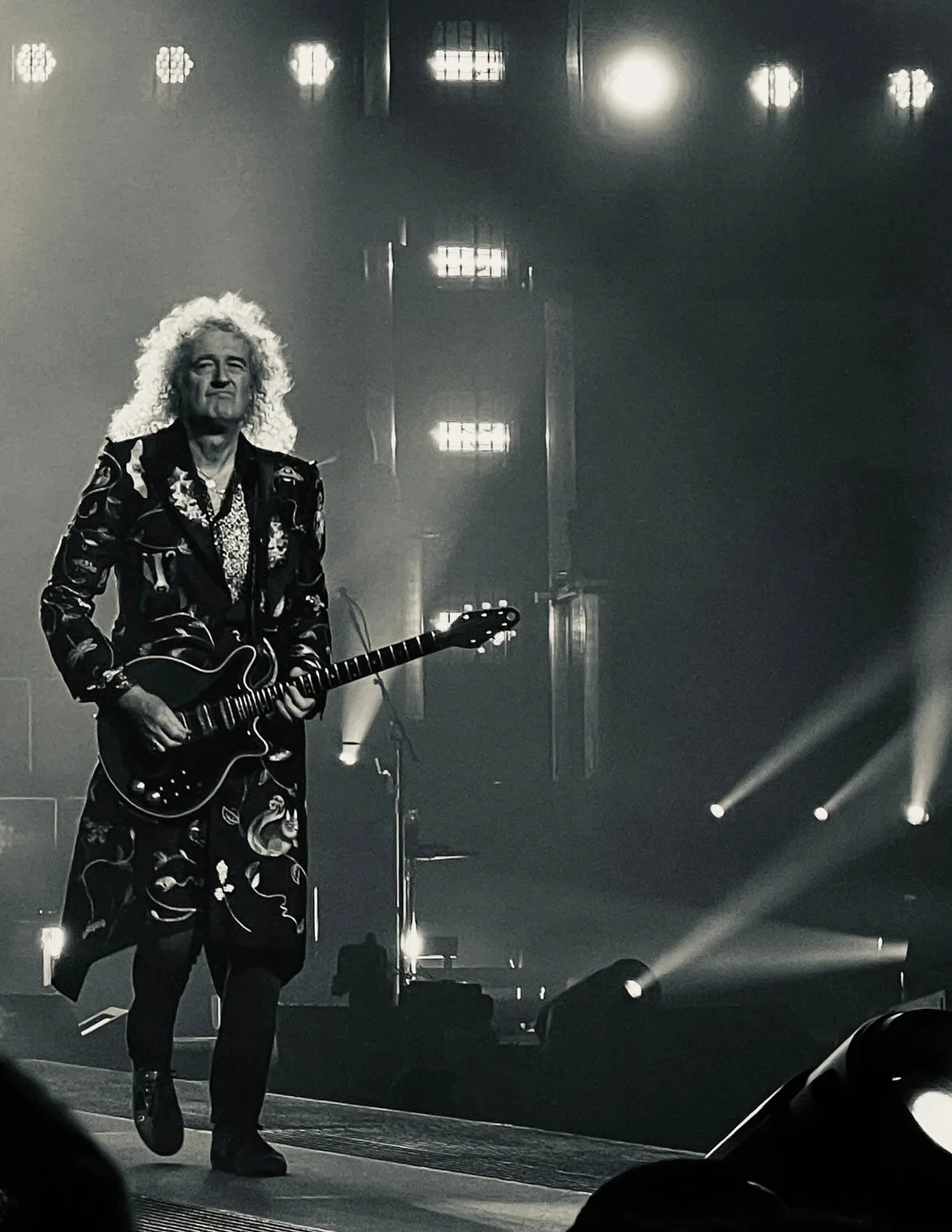 A black and white photo of a man with curly hair playing an electric guitar on stage with bright lights shining behind him.