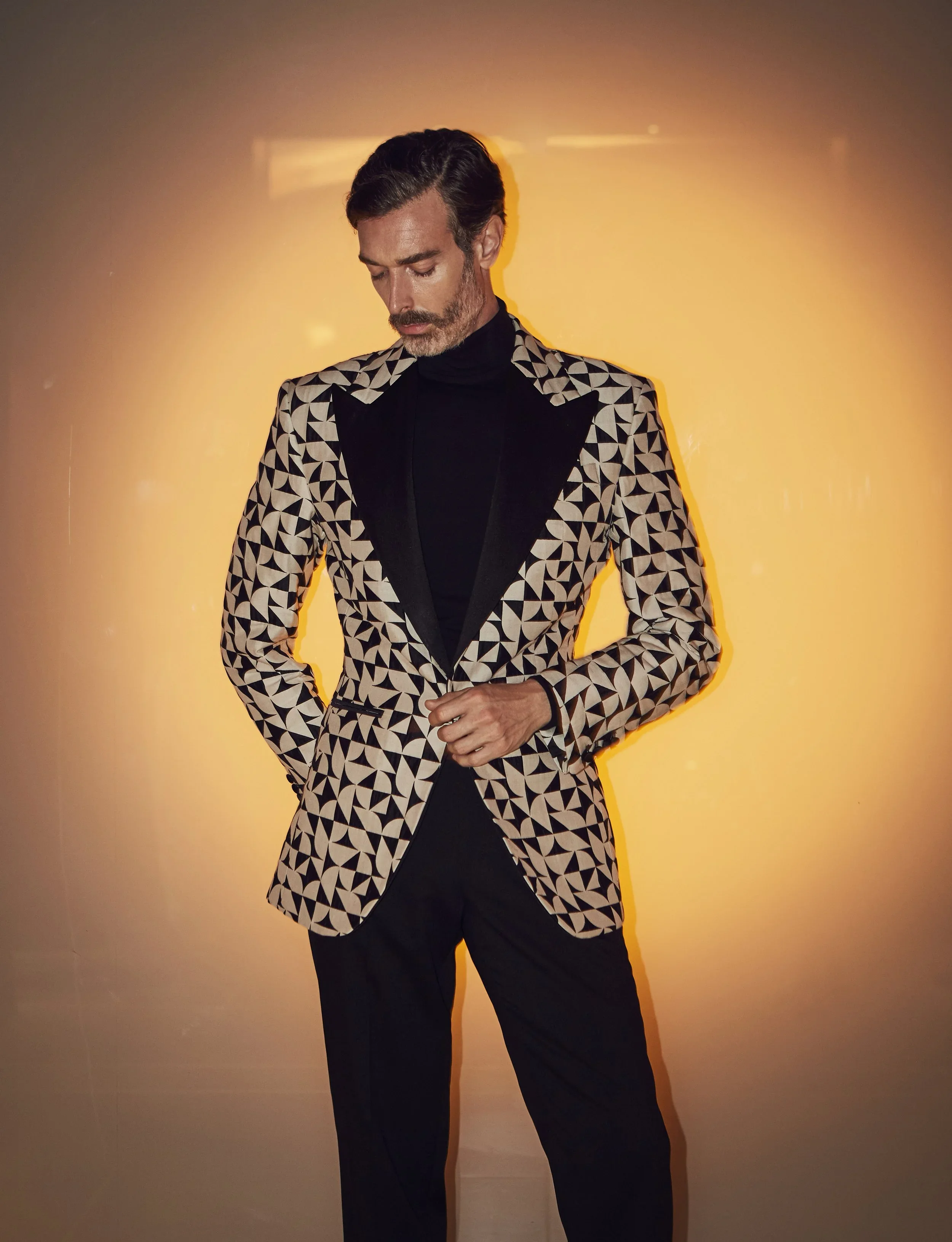 A man with dark hair and a beard wearing a black turtleneck and a patterned blazer with geometric shapes stands against a yellow-orange gradient background, looking downward with a serious expression.