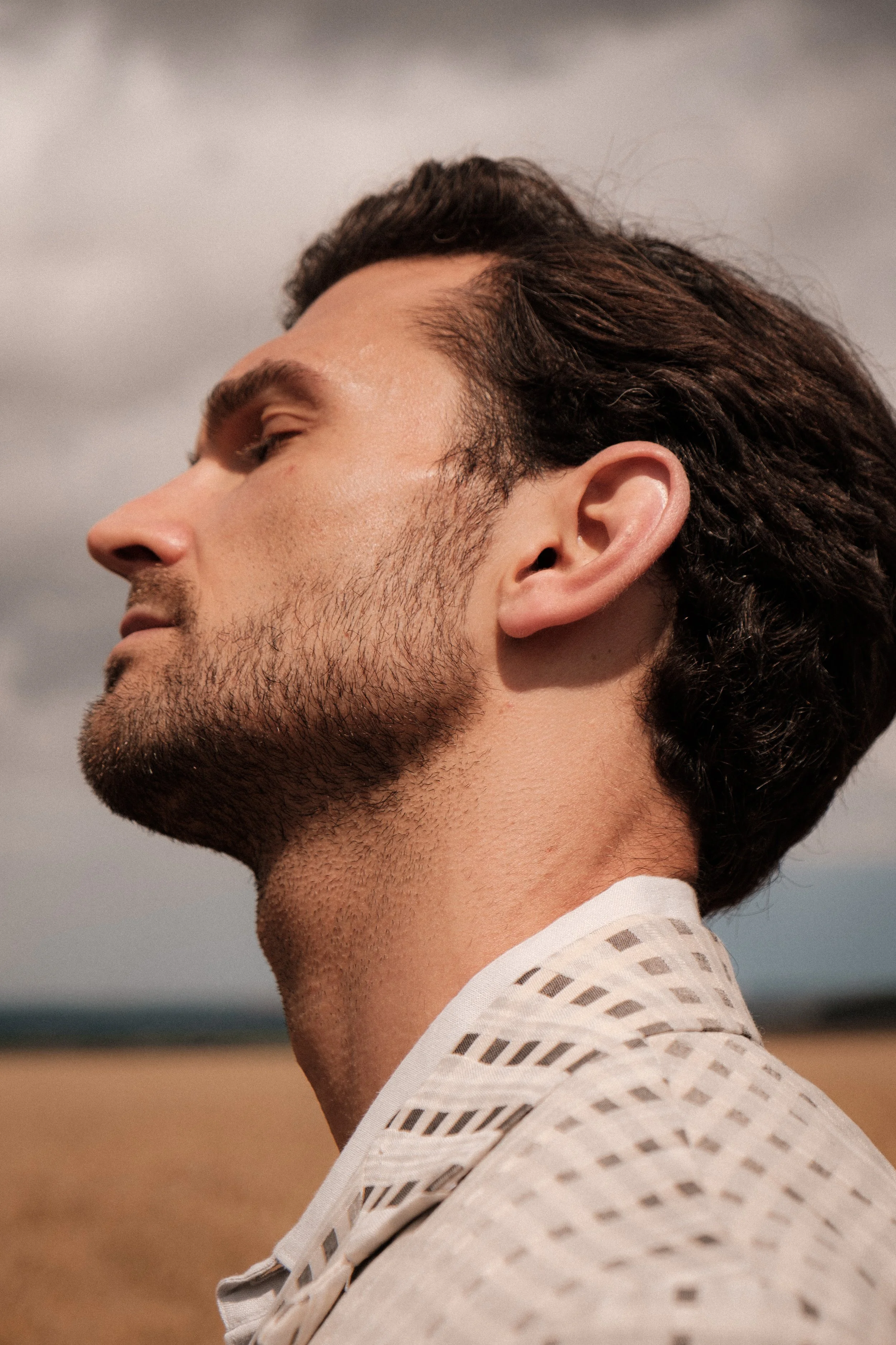 Close-up side profile of a young man with dark, wavy hair and light skin, wearing a white shirt with cut-out patterns, standing outdoors with a blurred landscape in the background.