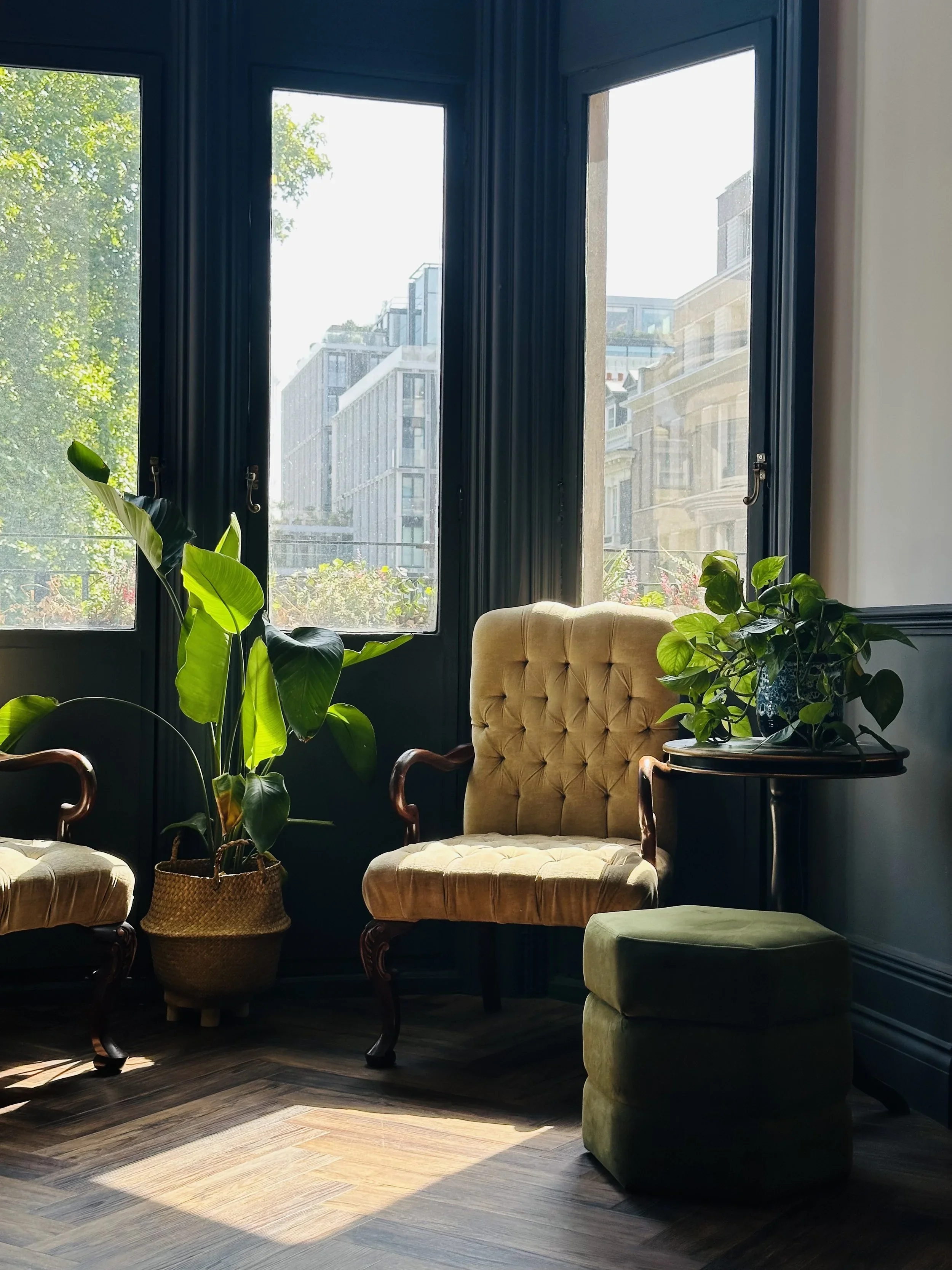 Living room with large windows, green plants, a tufted armchair, a small round side table, and patterned wood flooring illuminated by sunlight.