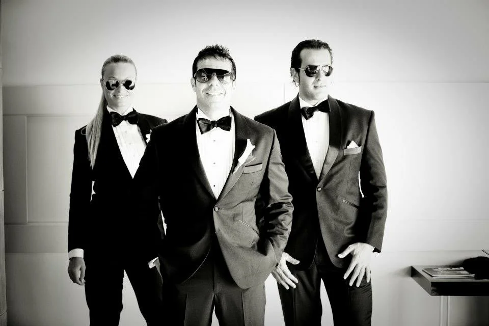 Three people dressed in tuxedos and sunglasses smiling and posing in a modern room.