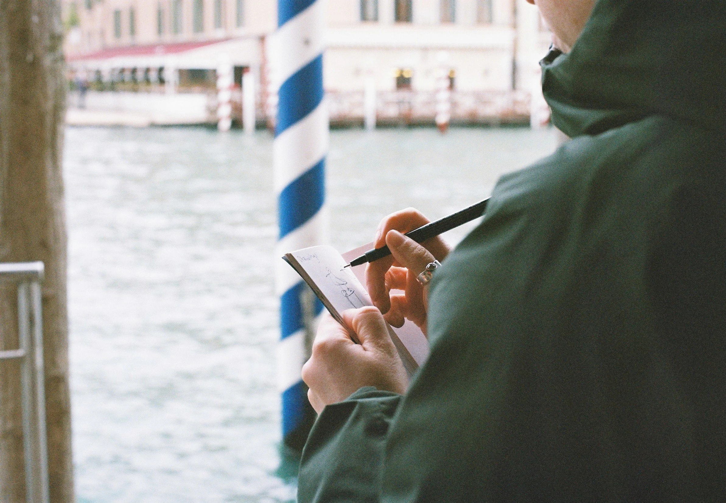 Person writing in a small notepad while standing near a canal with a striped pole in Venice, Italy.