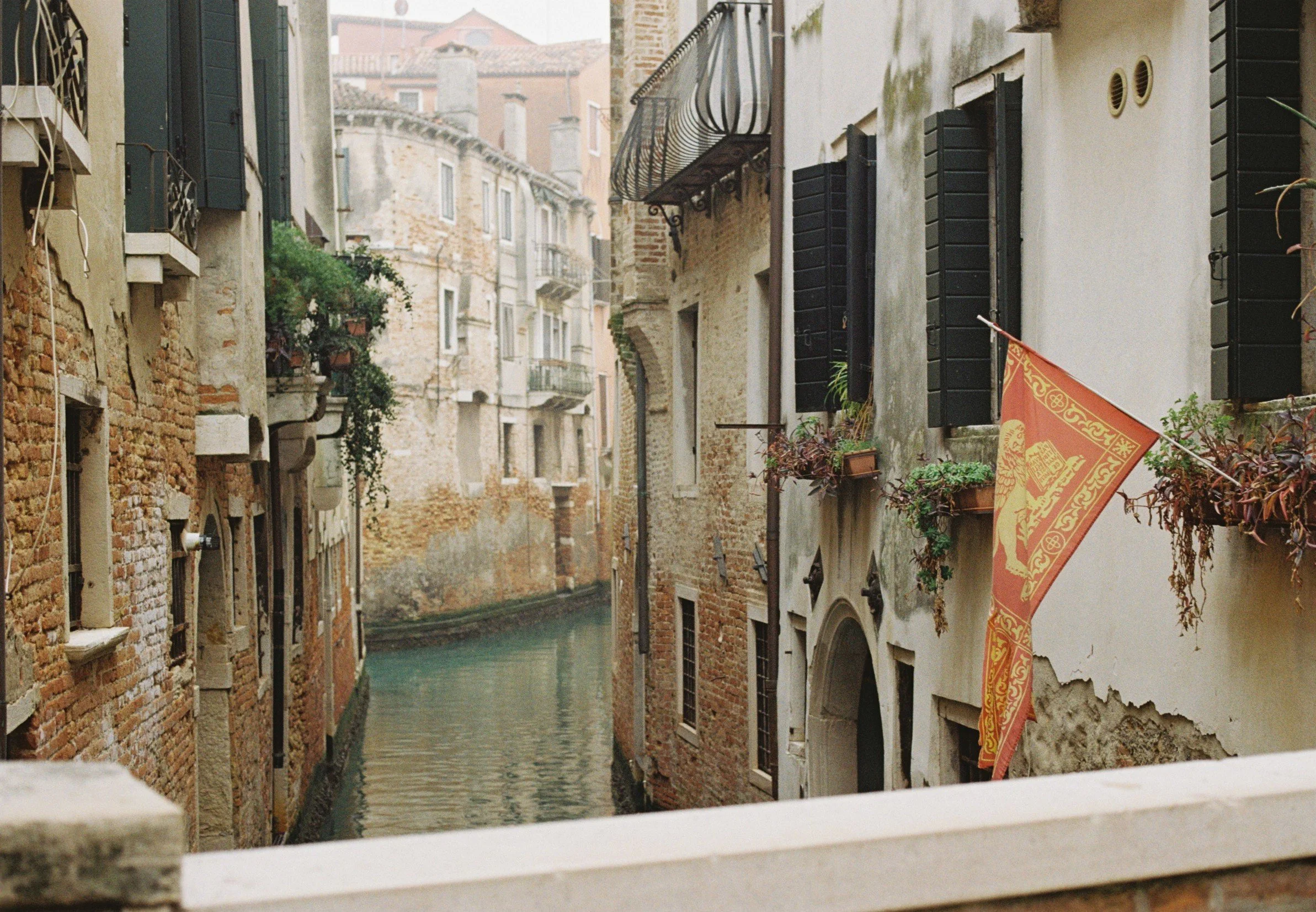 A view of a narrow canal through a small opening, with old buildings on both sides featuring shutters and balconies, in Venice.
