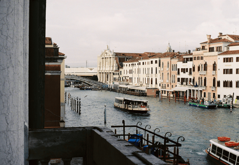 A canal scene in Venice, Italy, with boats on the water, historic buildings along the canal, and a bridge in the background.