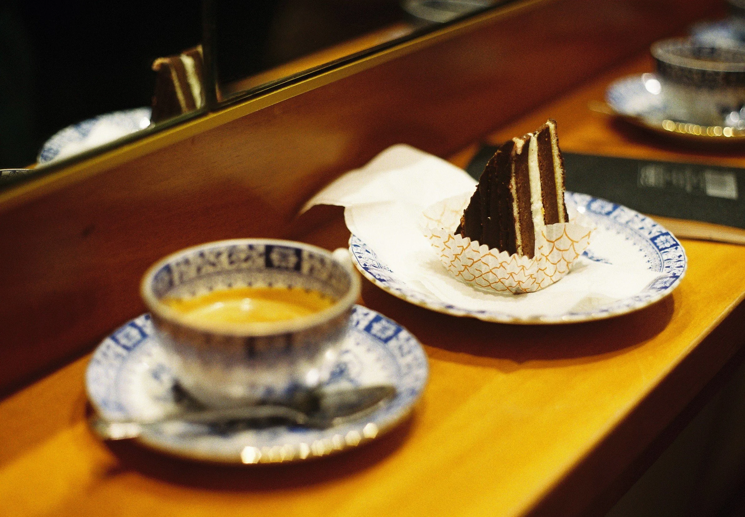 Chocolate cake with cream filling on a patterned plate, accompanied by a small cup of yellow sauce or custard, displayed on a wooden surface.