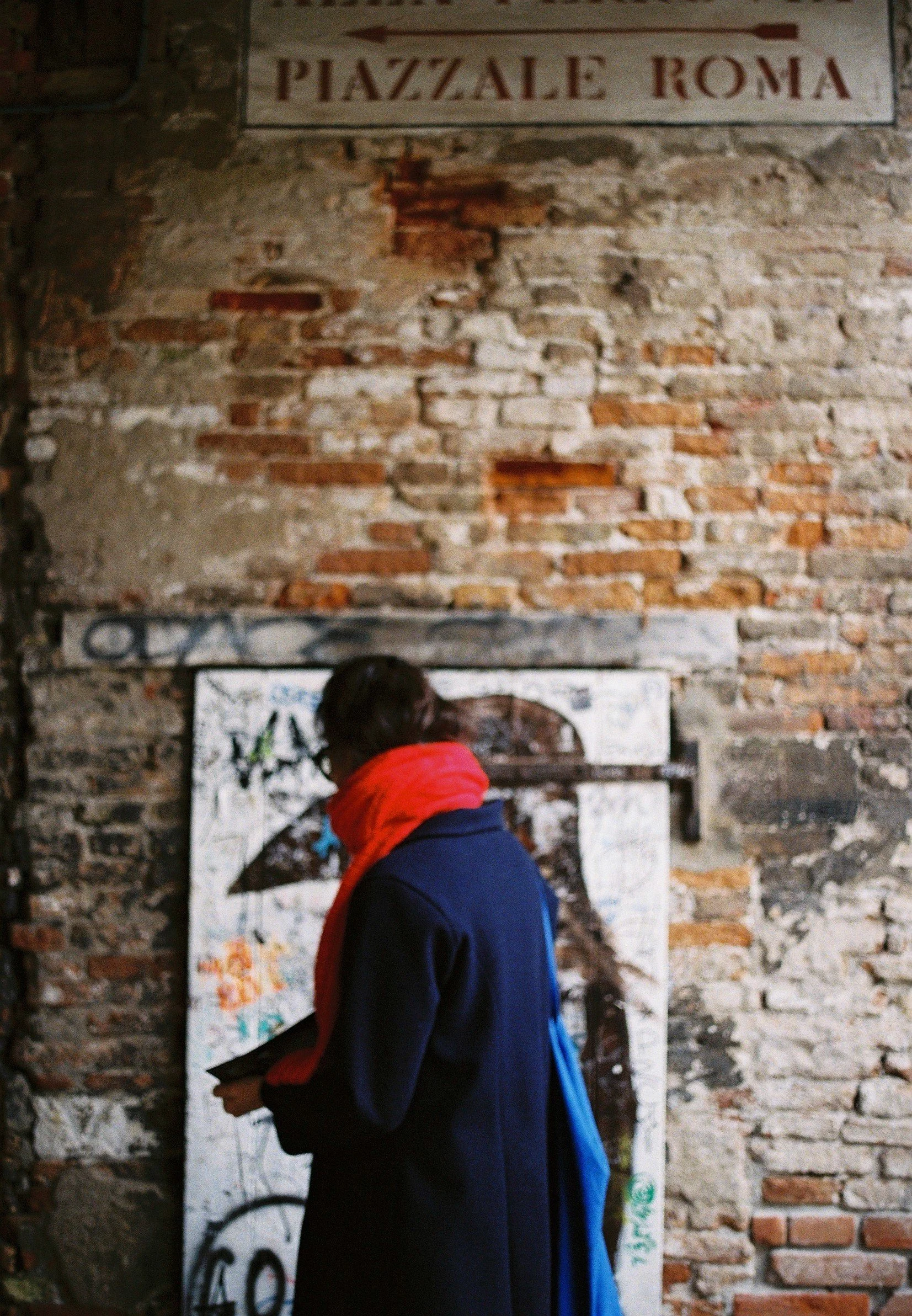 A person with dark hair wearing a blue coat and red scarf, standing in front of a brick wall with graffiti and street signs, reading a piece of paper or booklet.